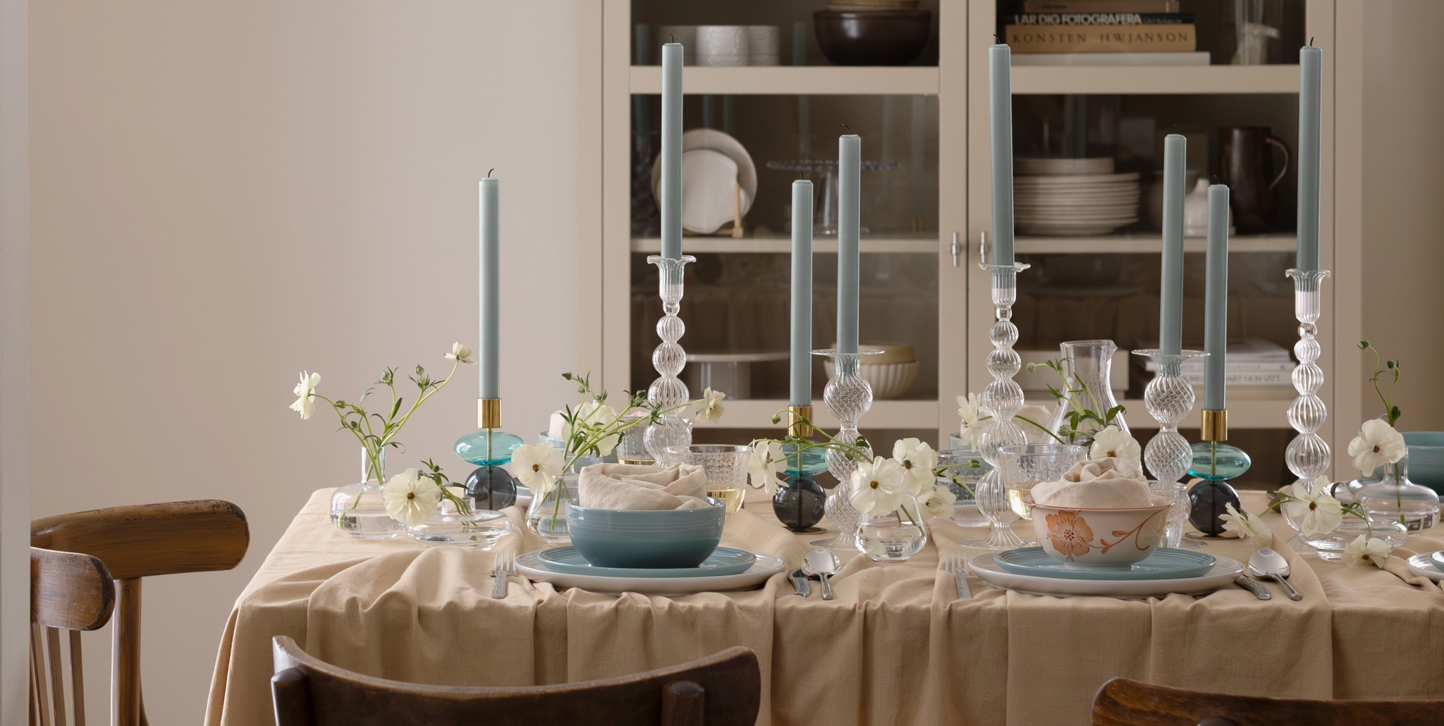 A dining table set with blue candles, white flowers, and dinnerware, next to a kitchen counter.
