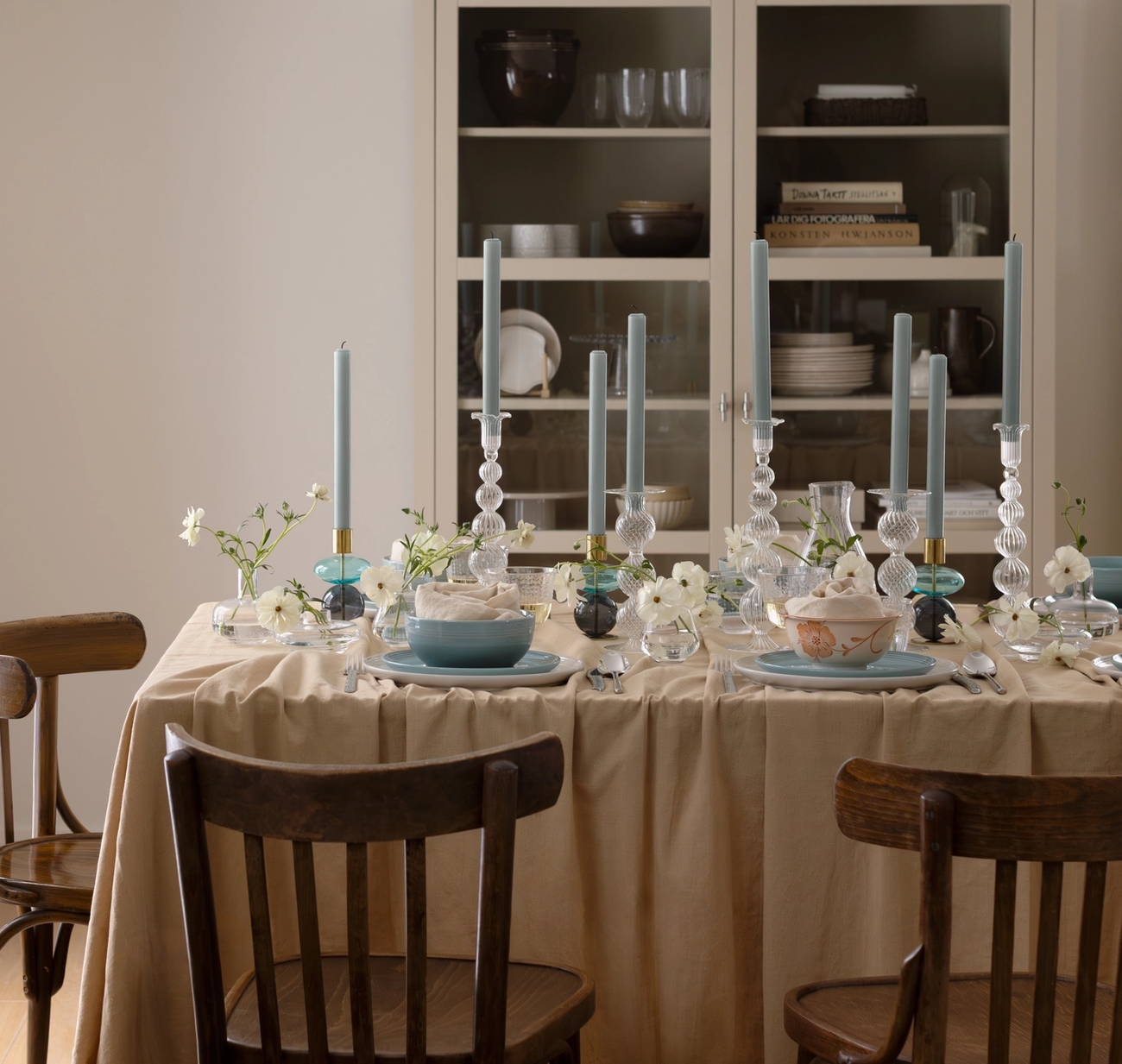 Ett matbord dukat med blå ljus, vita blommor och servis, bredvid en köksbänk med en kastrull.