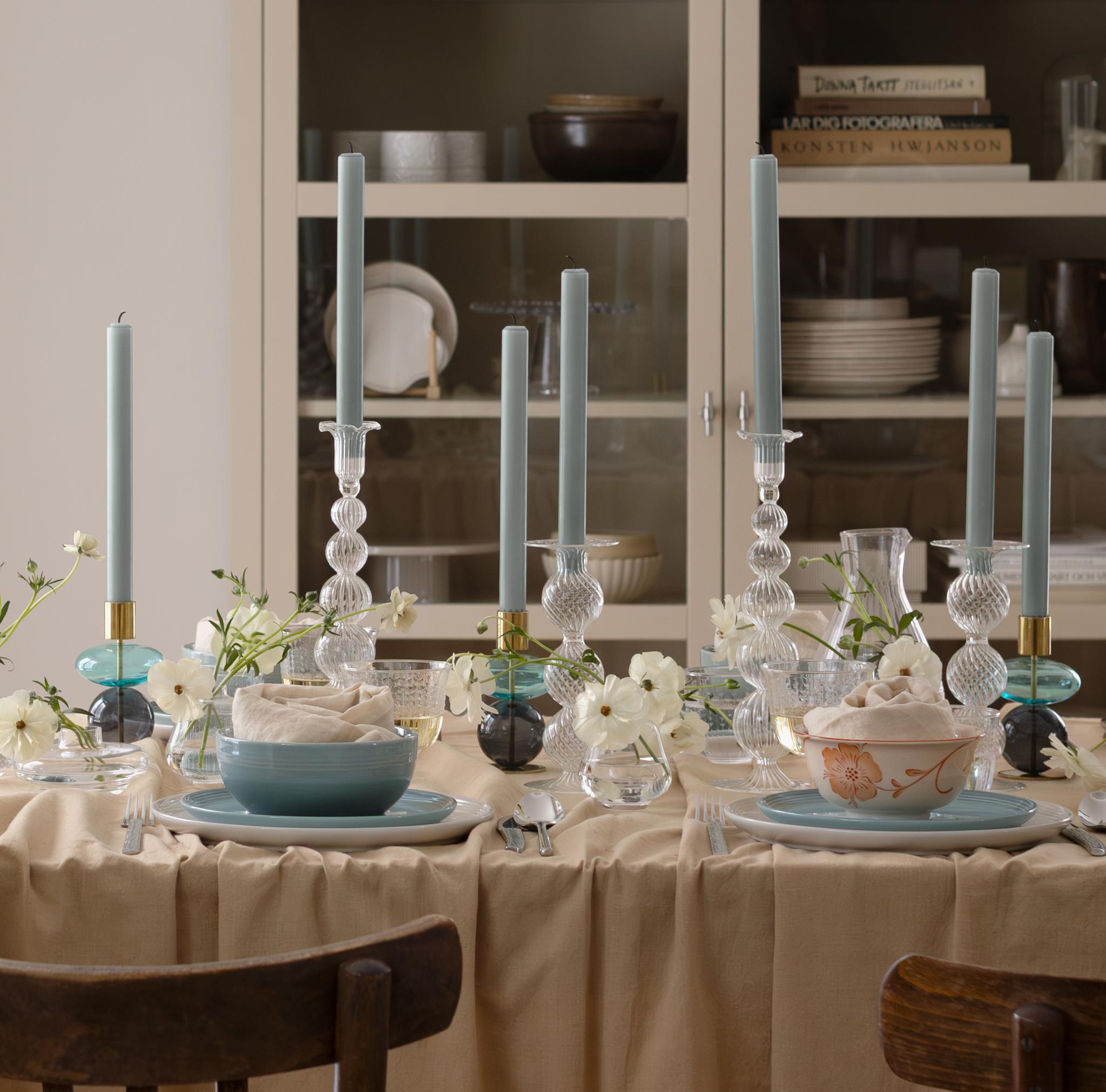 A dining table set with blue candles, white flowers, and dinnerware on a beige cloth, surrounded by wooden chairs, a kitchen counter, and a display cabinet.