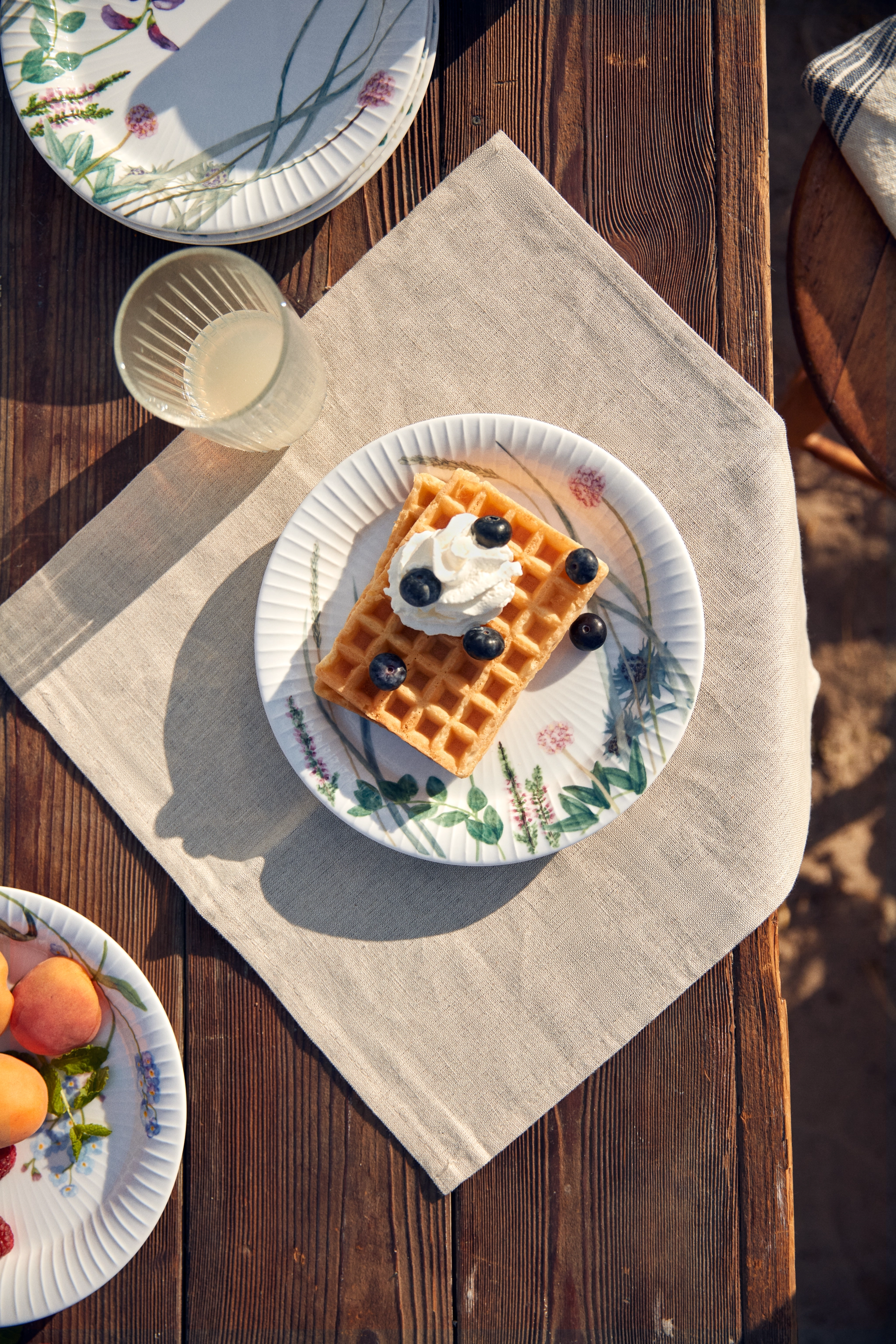 En våffla med vispad grädde och blåbär på en blommig tallrik, på en beige servett och ett träbord.