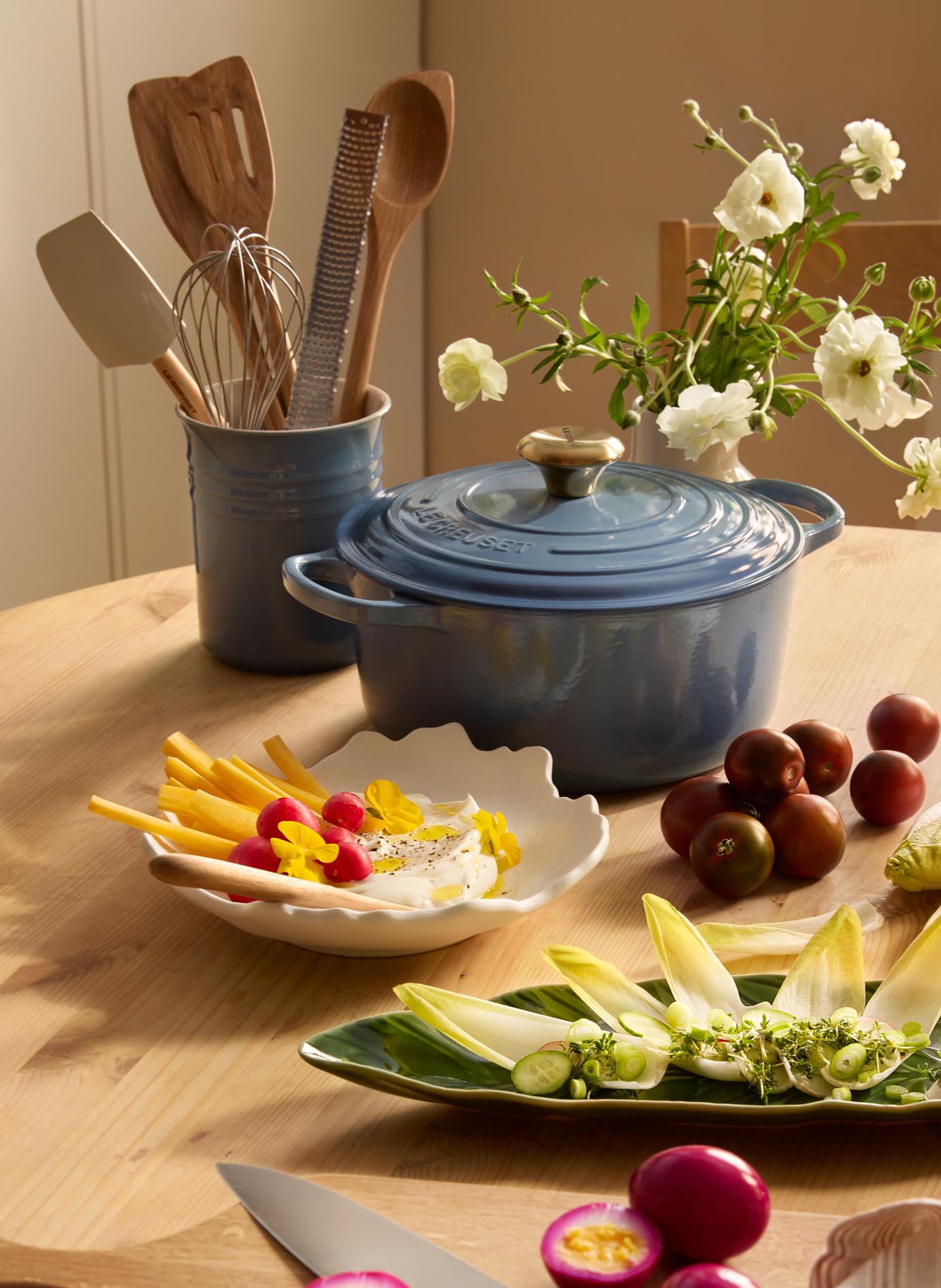 Sininen Le Creuset -pata puupöydällä, jota ympäröivät keittiövälineet, valkoiset kukat, kermainen dippi vihannesten kanssa, kirsikkatomaatit, endiivisalaatti ja viipaloidut pikkelöidyt kananmunat