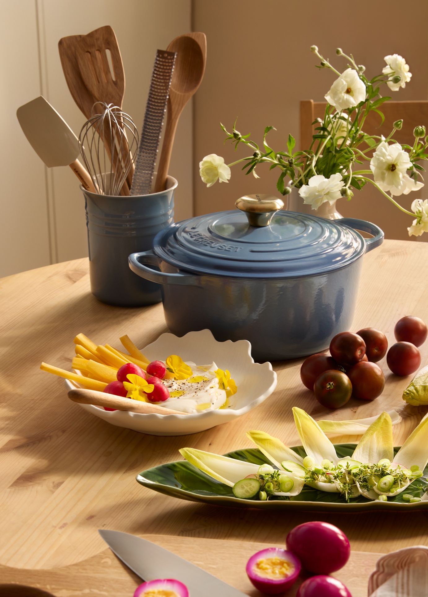 Sininen Le Creuset -pata puupöydällä, jota ympäröivät keittiövälineet, valkoiset kukat, kermainen dippi vihannesten kanssa, kirsikkatomaatit, endiivisalaatti ja viipaloidut pikkelöidyt kananmunat