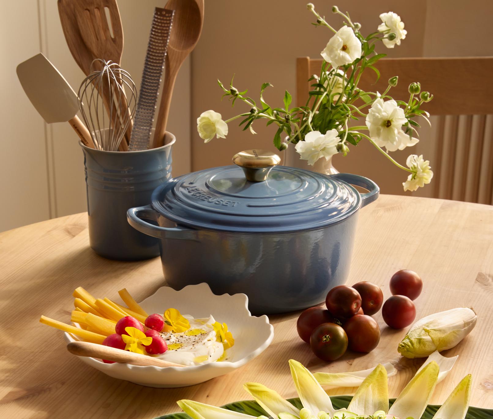 Sininen Le Creuset -ruukku ja erilaisia tuoreita ruokia, kuten punajuurella pikkelöityjä munia ja endiiviä, aseteltuna puupöydälle.