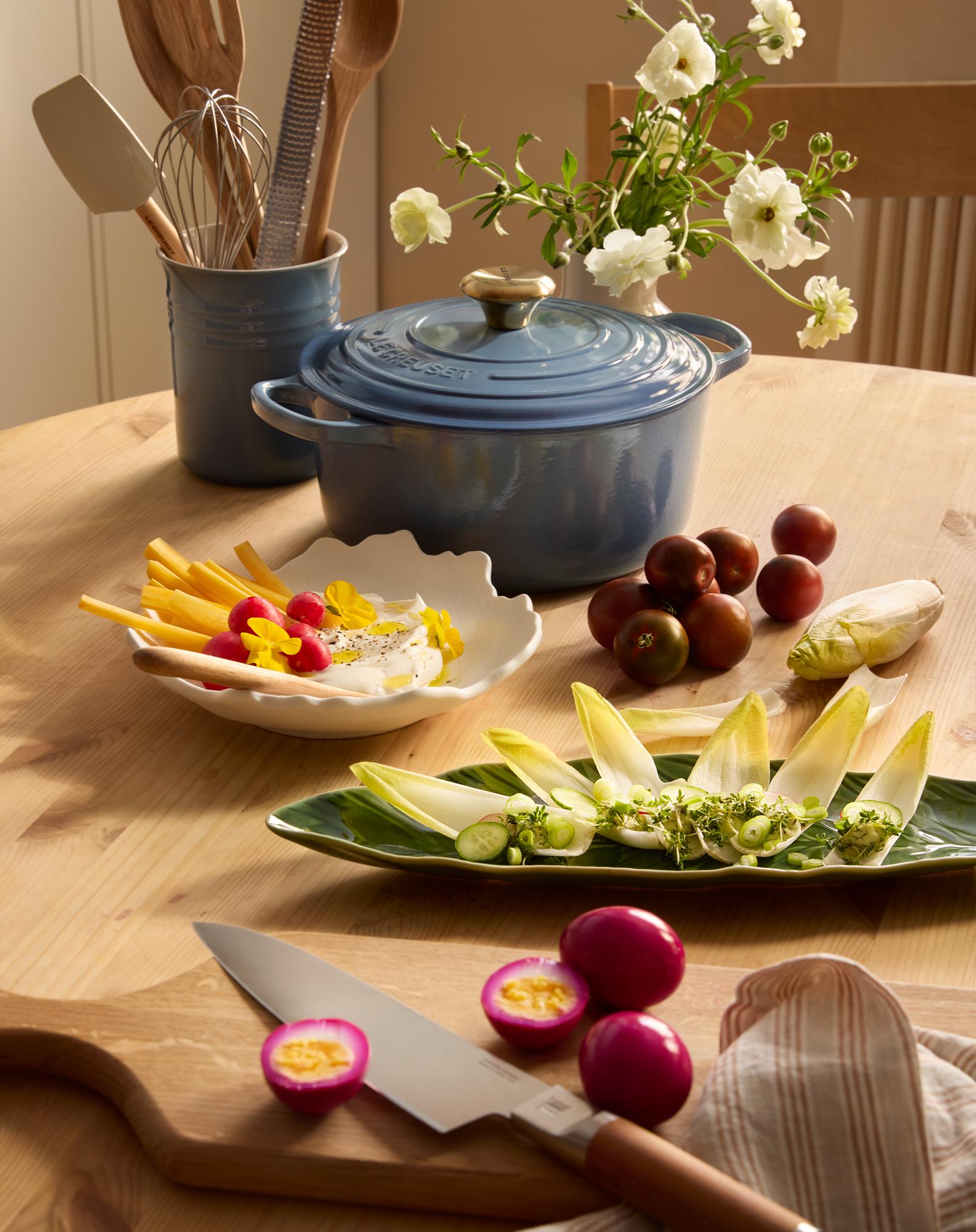 En blå Le Creuset-gryta, ett redskapskrus, vita blommor, olika förberedda snacks och magentafärgade inlagda ägg är arrangerade på ett träbord