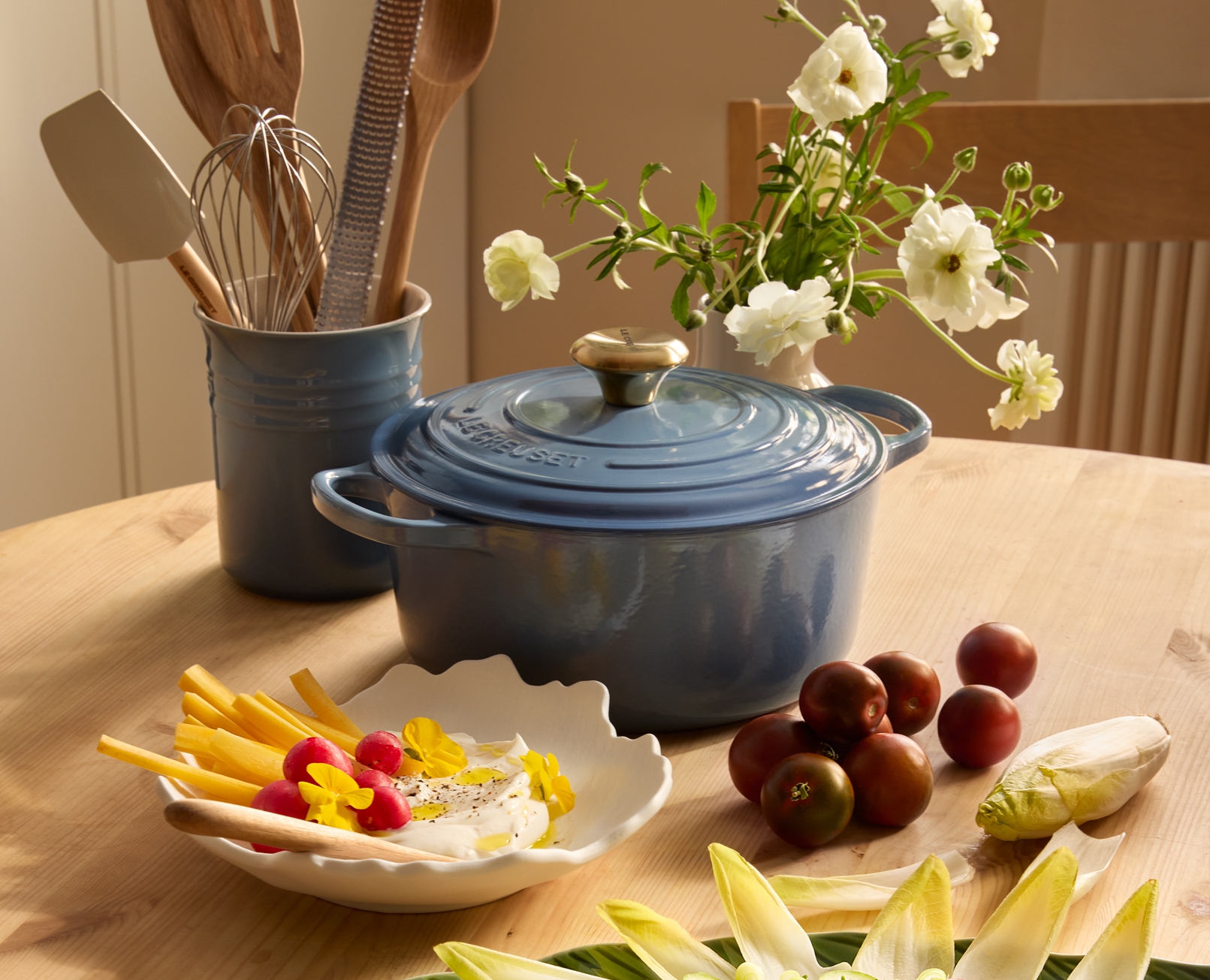 Sininen Le Creuset -ruukku ja erilaisia tuoreita ruokia, kuten punajuurella pikkelöityjä munia ja endiiviä, aseteltuna puupöydälle.