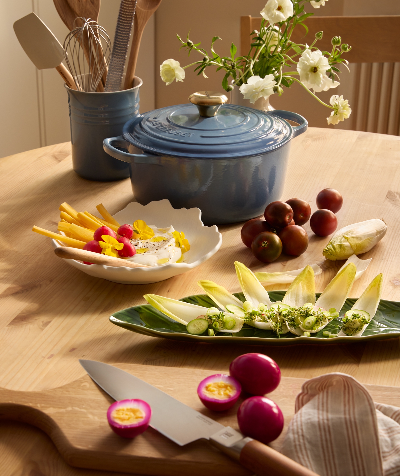 En blå Le Creuset-gryta, ett redskapskrus, vita blommor, olika förberedda snacks och magentafärgade inlagda ägg är arrangerade på ett träbord