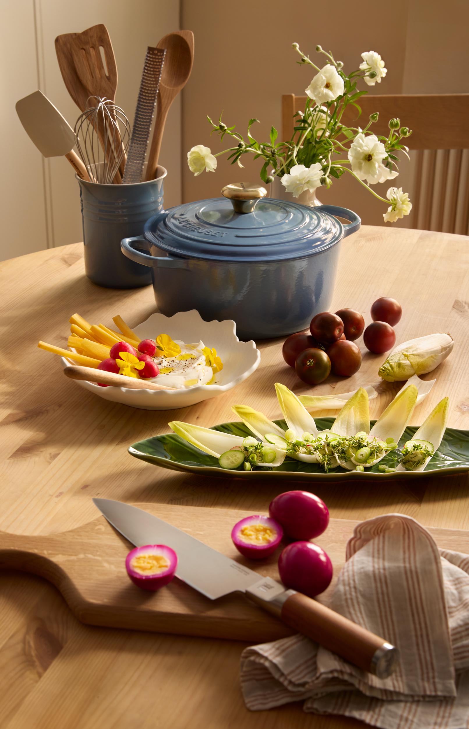 En blå Le Creuset-gryte står på et trebord omgitt av kjøkkenredskaper, hvite blomster, en dipp med reddiker og gulrøtter, en endiviesalat, tomater og syltede egg