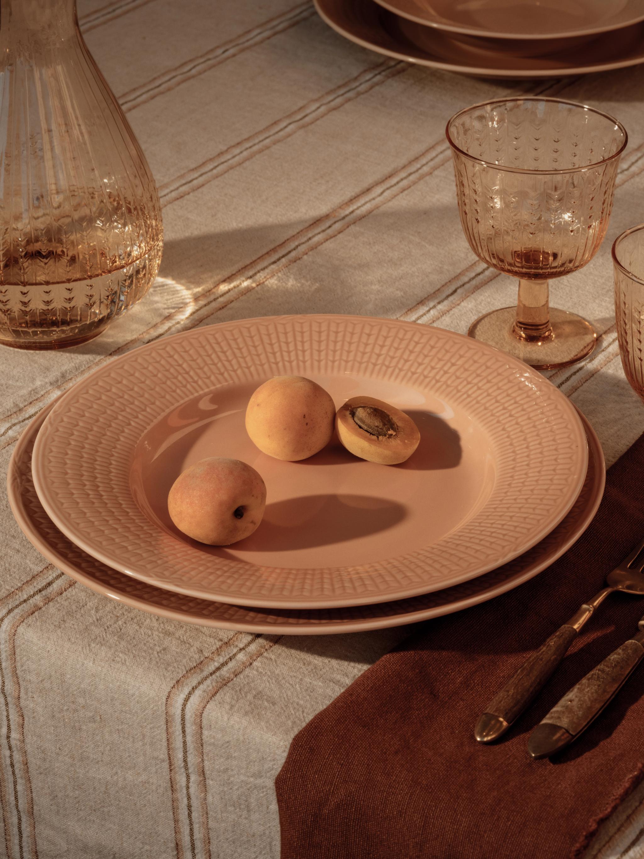 Et spisebord dekket med karamellfargede tallerkener med aprikoser, en glasskanne, et teksturert glass og bestikk på en brun serviett, på en stripete duk.