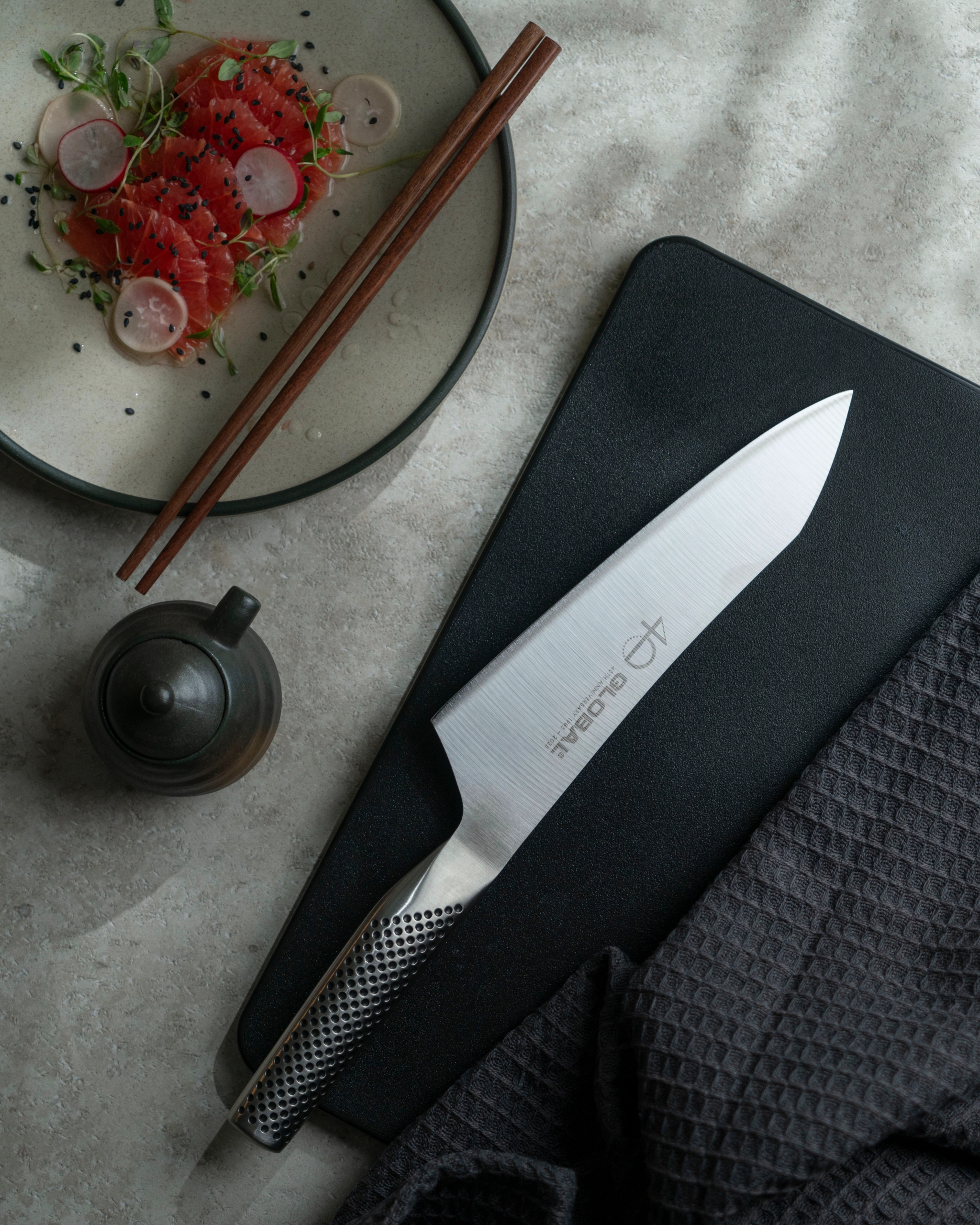 Ylhäältäpäin otettu kuva näyttää Global-kokkiveitsen mustalla leikkuulaudalla, lautasen sashimia syömäpuikkoineen, pienen kattilan ja tummanharmaa pyyhe harmaalla työtasolla.