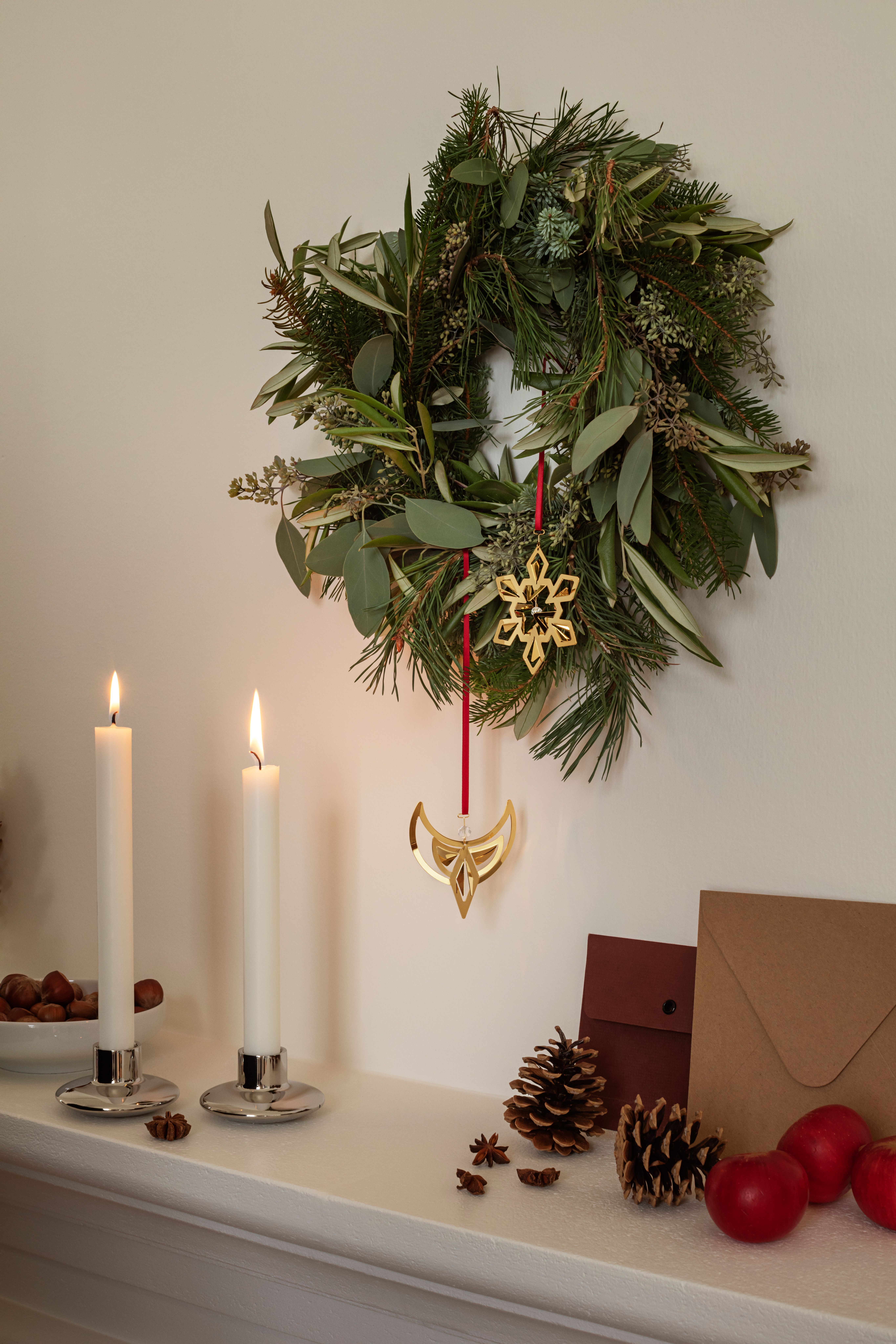 Grön julkrans med guldfärgade hängen ovanför tända ljus, kottar och röda julkulor.