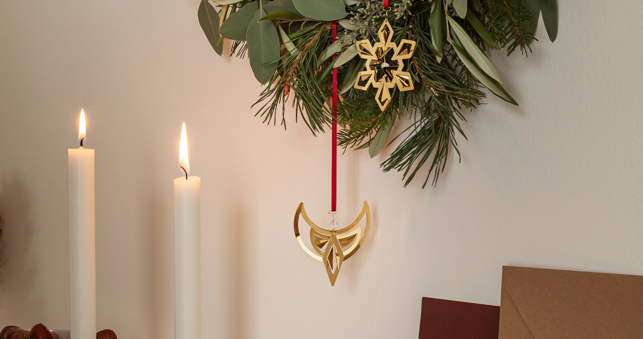 Grön julkrans med guldfärgade hängen ovanför tända ljus, kottar och röda julkulor.
