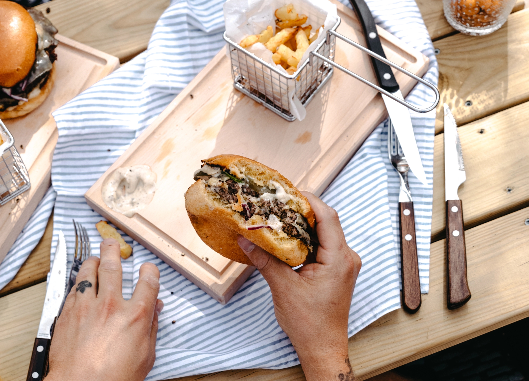 Person med tatueringar äter en hamburgare med pommes frites vid ett träbord utomhus.