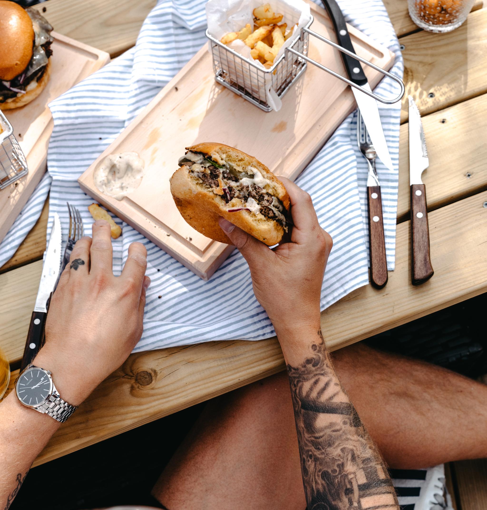 Person med tatueringar äter en hamburgare vid ett träbord utomhus med pommes frites och bestick runtomkring.