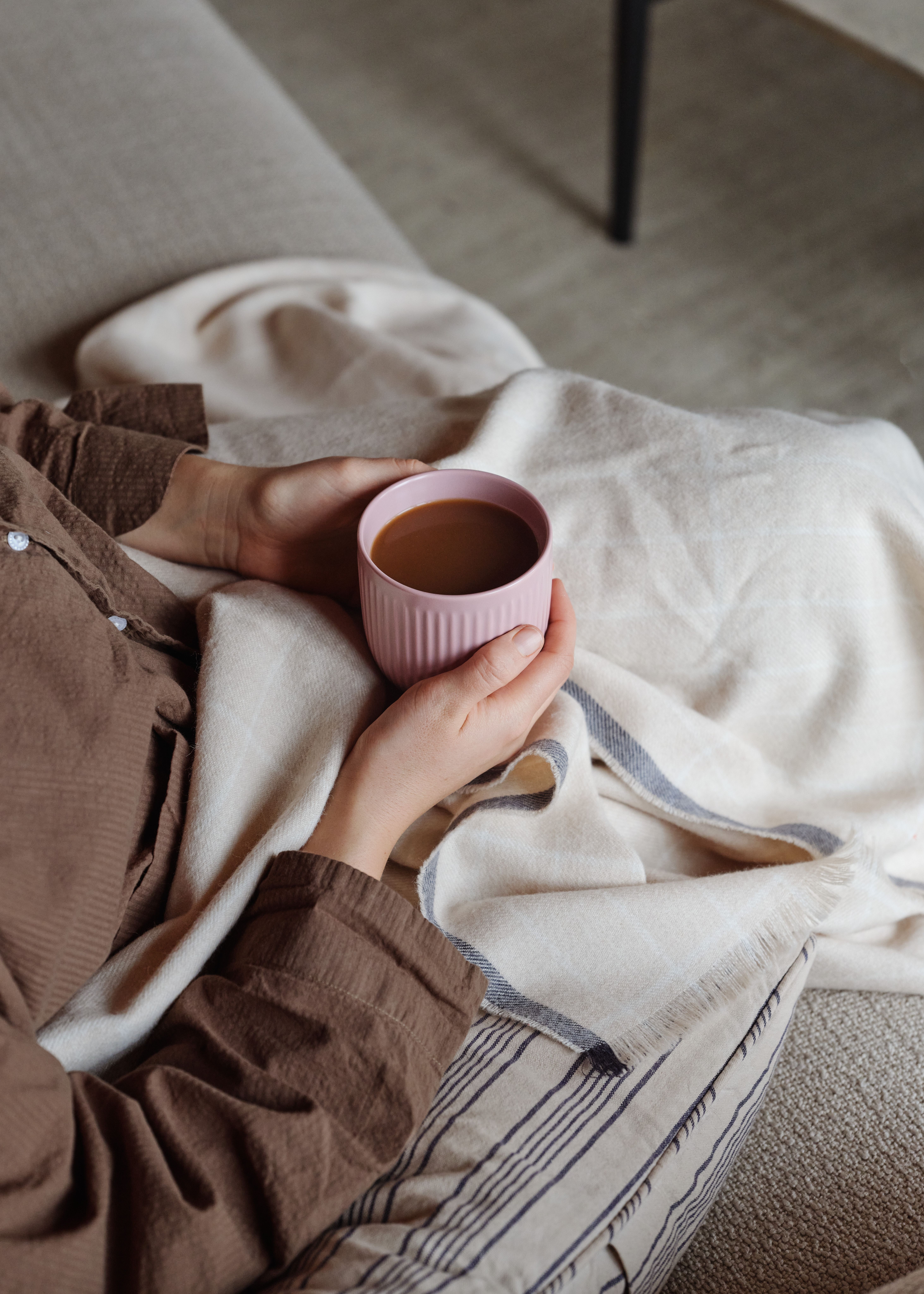 Kädet pitelevät vaaleanpunaista kahvimukia mukavasti viltin alla.