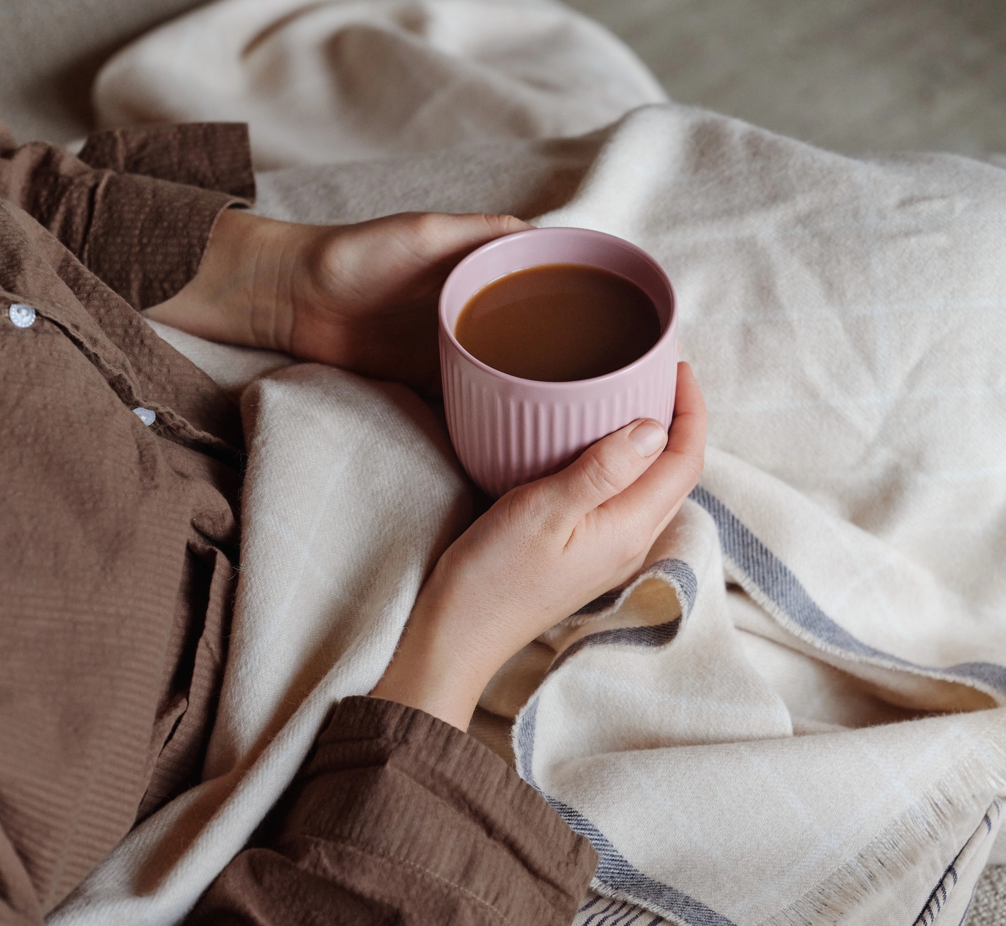 Kädet pitelevät vaaleanpunaista kahvimukia mukavasti viltin alla.