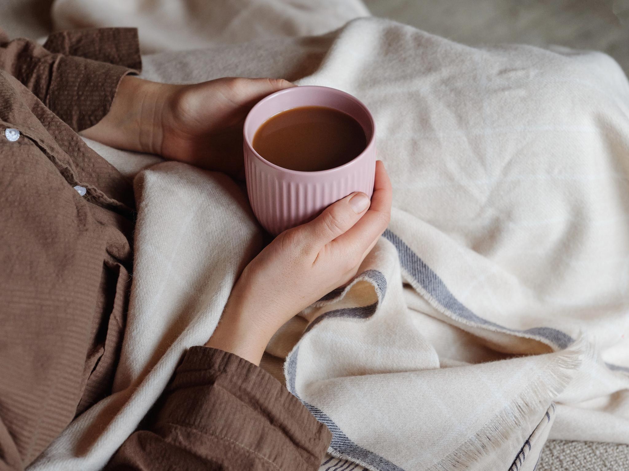 Kädet pitelevät vaaleanpunaista kahvimukia mukavasti viltin alla.