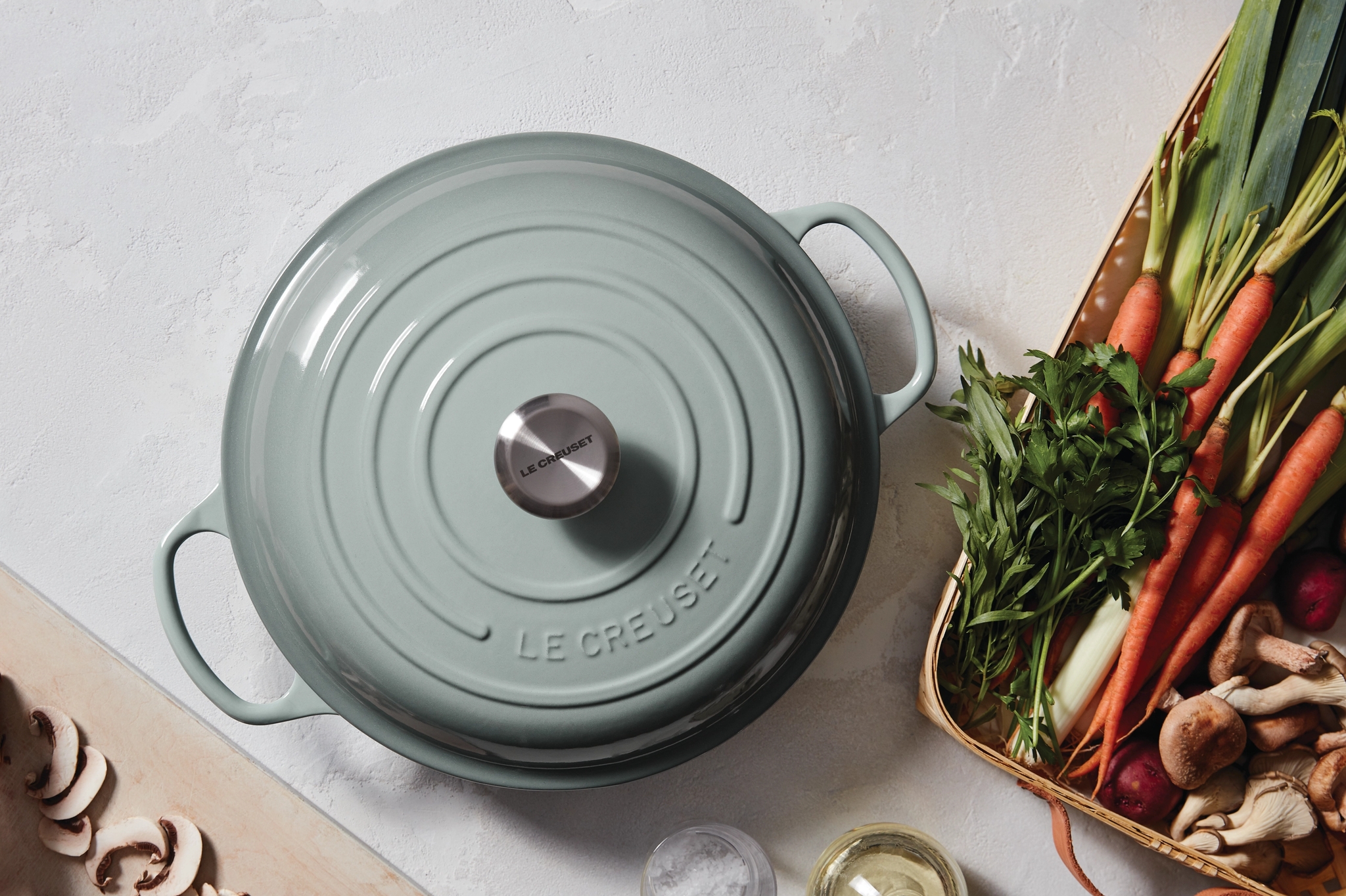 Vaaleanvihreä Le Creuset -pata keittiötasolla tuoreiden vihannesten ja sienien kanssa ruoanlaittoa varten.