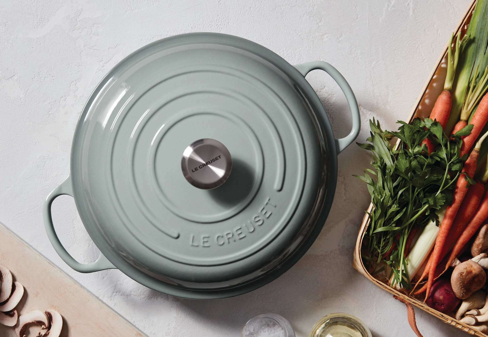 Vaaleanvihreä Le Creuset -pata keittiötasolla tuoreiden vihannesten ja sienien kanssa ruoanlaittoa varten.