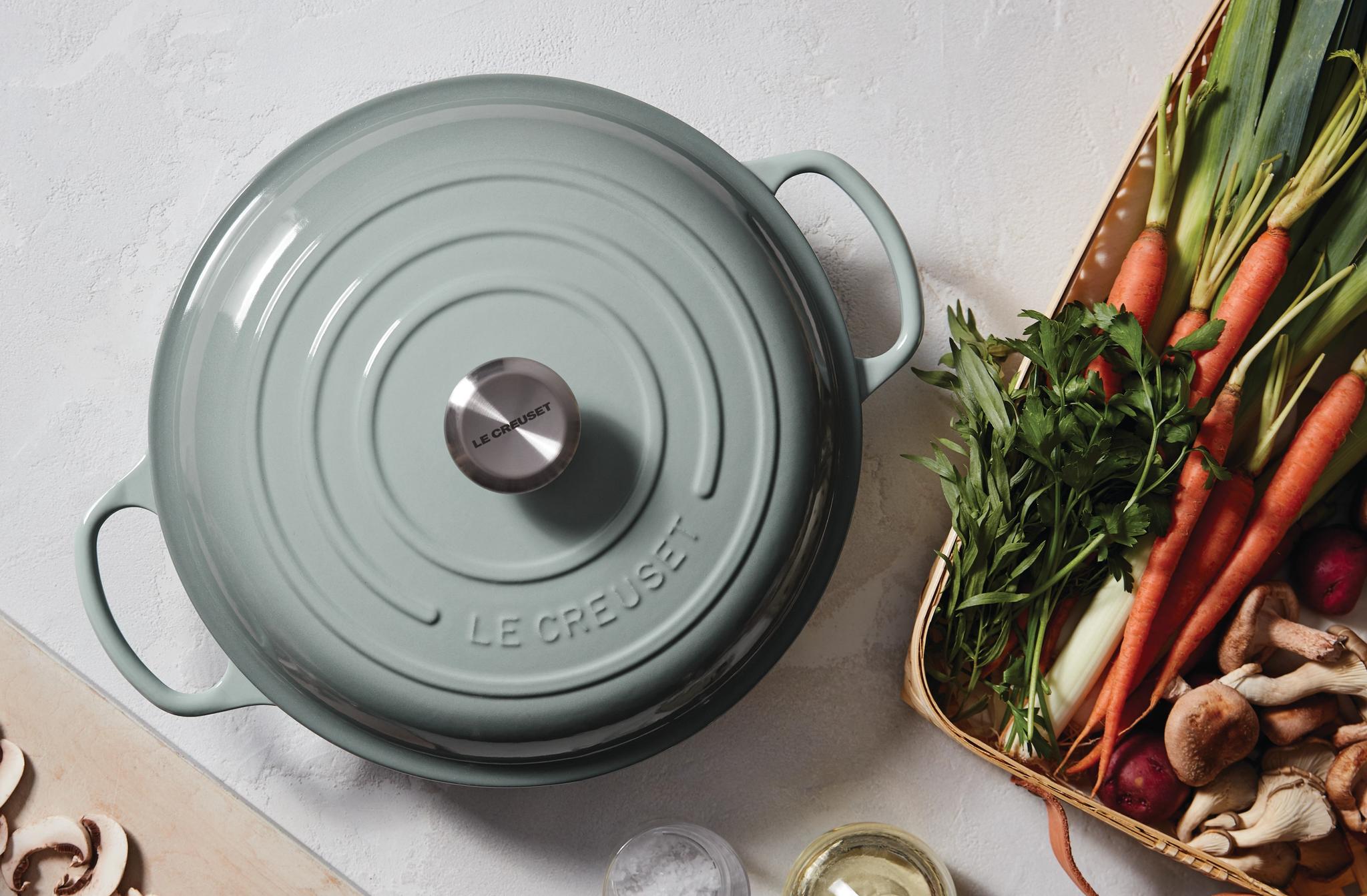 Vaaleanvihreä Le Creuset -pata keittiötasolla tuoreiden vihannesten ja sienien kanssa, kun kädet viipaloivat sieniä leikkuulaudalla.