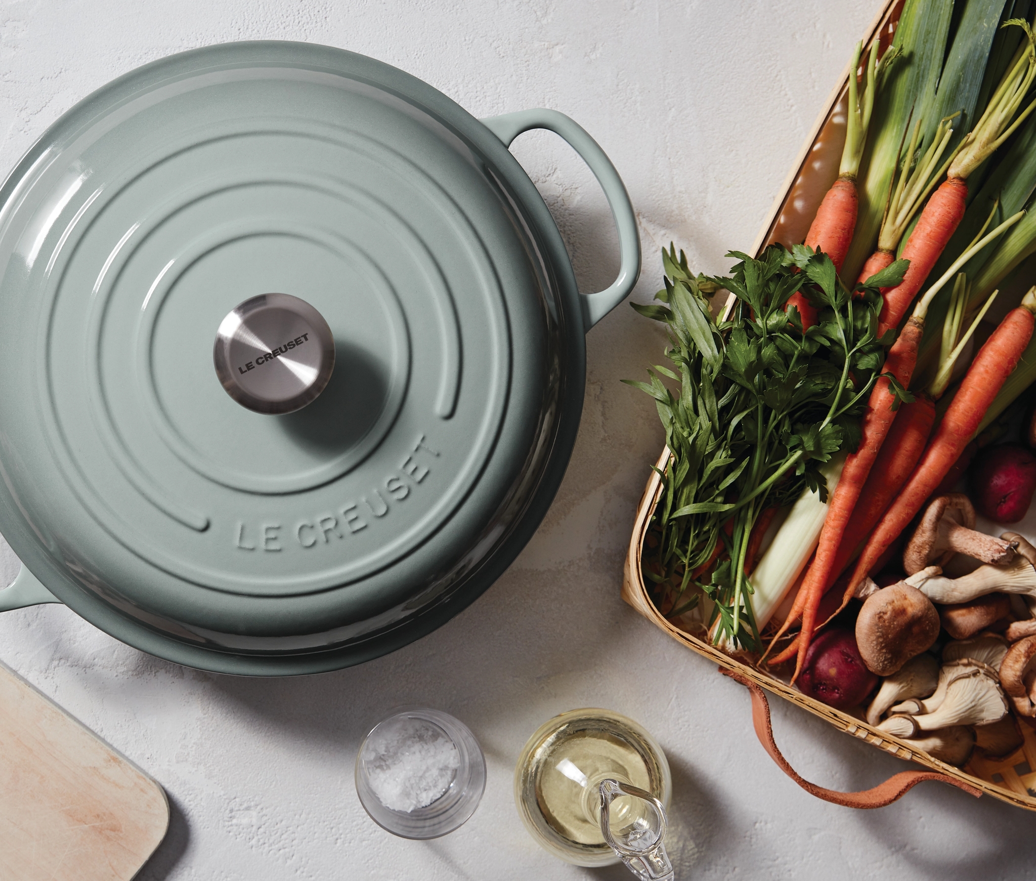 Ylhäältä katsottuna vaaleanvihreä Le Creuset -pata, sieniä viipaloivia käsiä ja kori tuoreita vihanneksia, kuten porkkanoita ja purjoa.