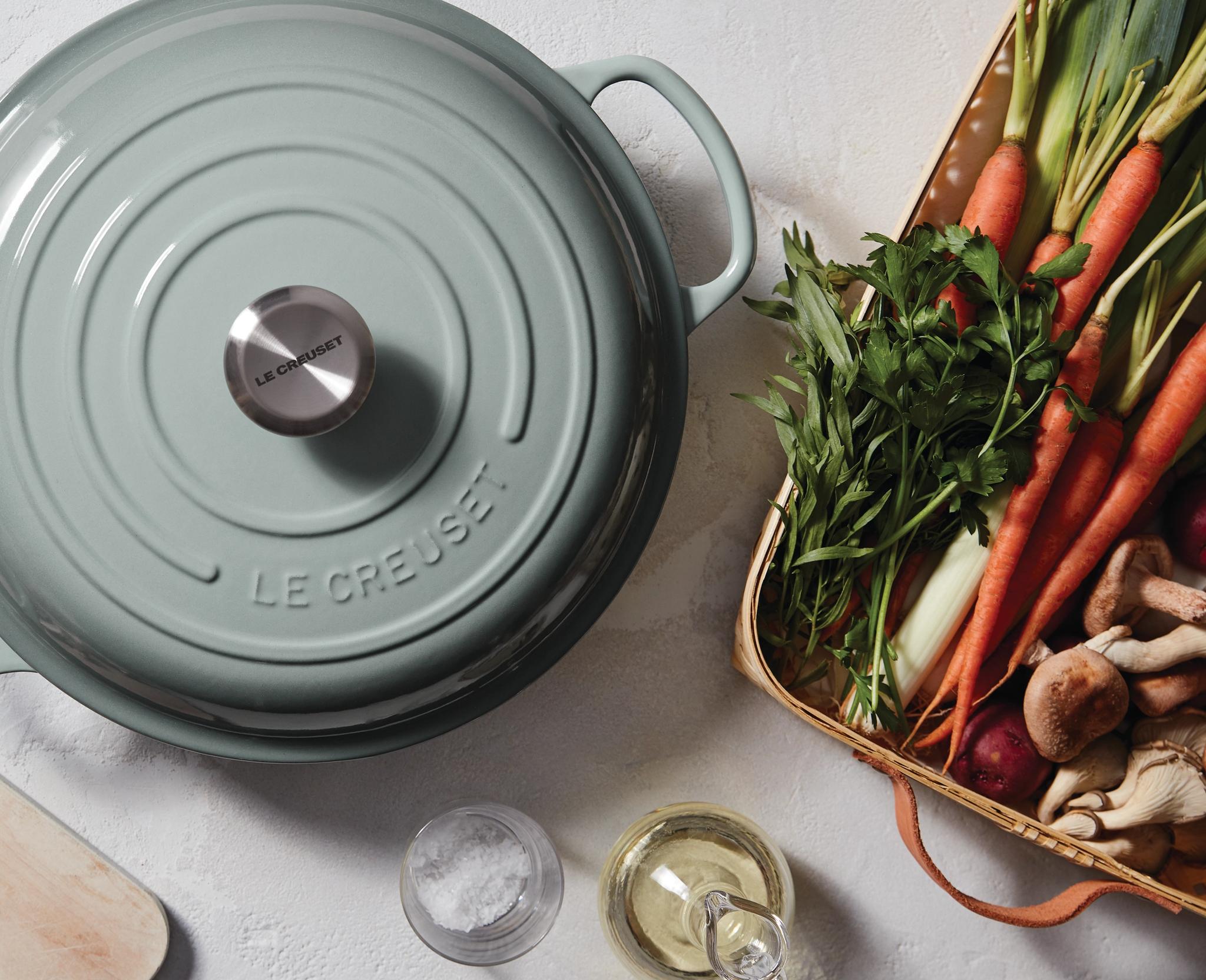 Ylhäältä katsottuna vaaleanvihreä Le Creuset -pata, sieniä viipaloivia käsiä ja kori tuoreita vihanneksia, kuten porkkanoita ja purjoa.