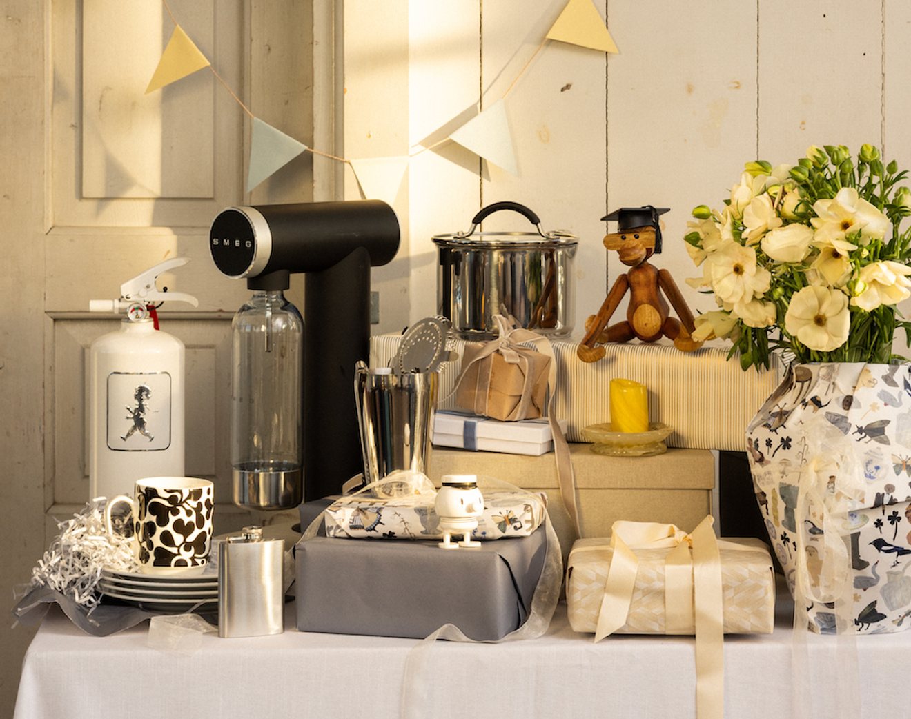 A table full of gifts for a celebration, including a black Smeg soda maker, a white fire extinguisher, a wooden graduation doll, and a vase of white flowers, with bunting flags above.