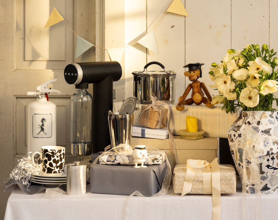 A table full of gifts for a celebration, including a black Smeg soda maker, a white fire extinguisher, a wooden graduation doll, and a vase of white flowers, with bunting flags above.