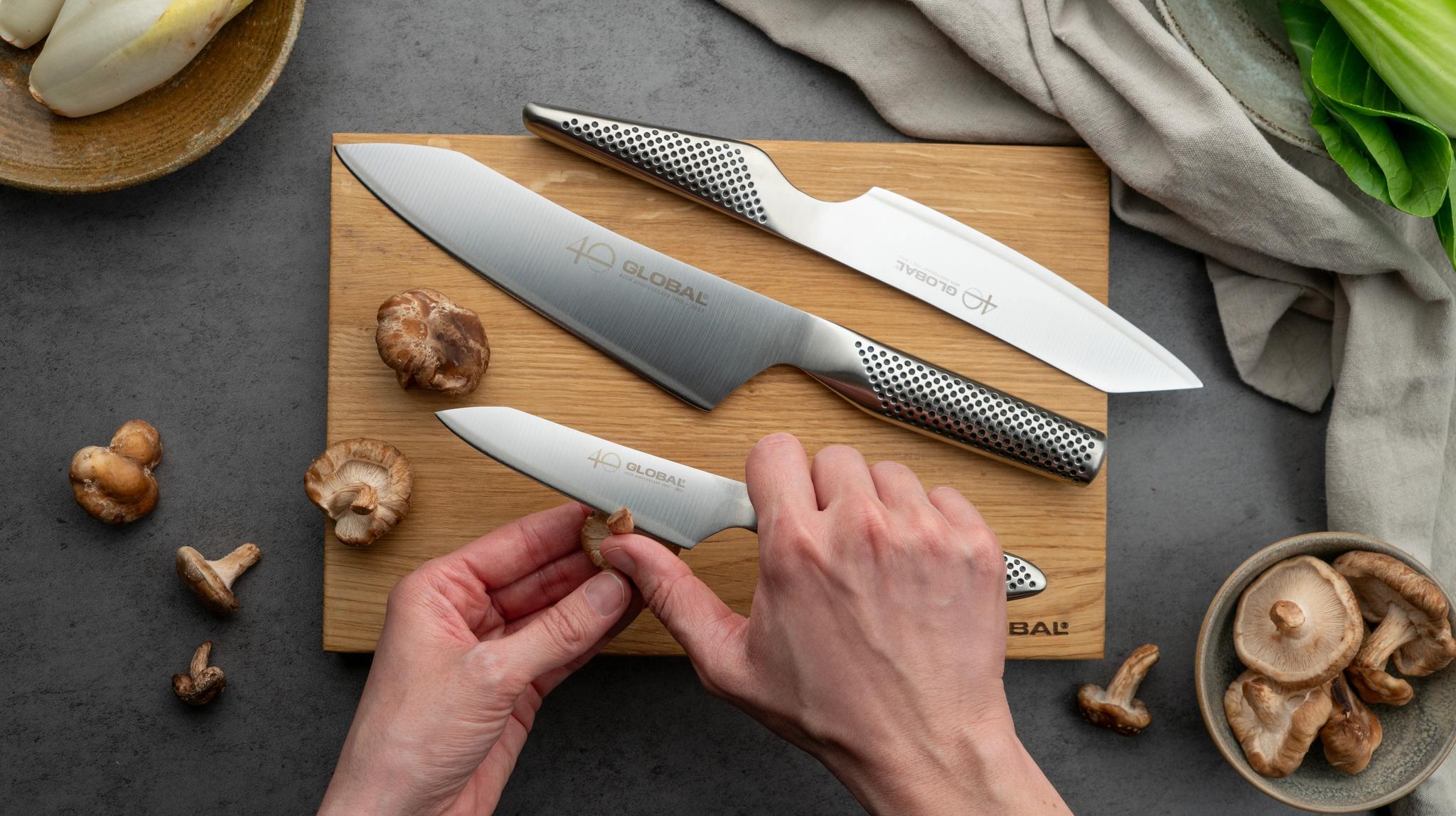 Henkilö kuorii sienen leikkuulaudalla Global-veitsellä muiden Global-veitsien ja tuoreiden tuotteiden ympäröimänä.