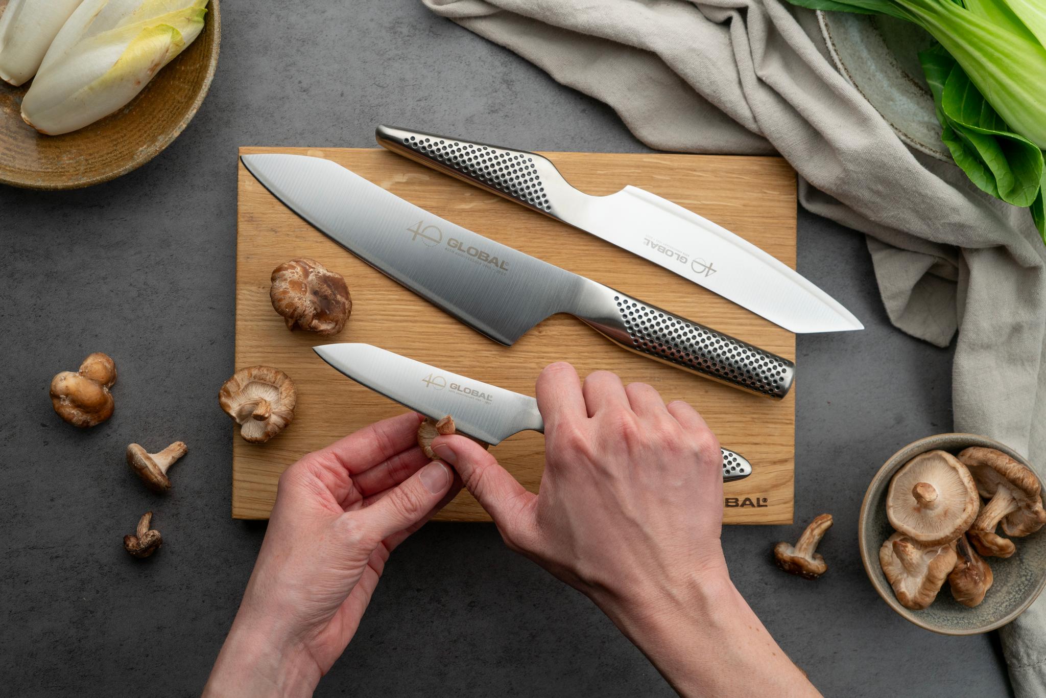 Händer som trimmar en shiitakesvamps stjälk med en Global-kniv, med andra Global-knivar och svampar på en skärbräda.