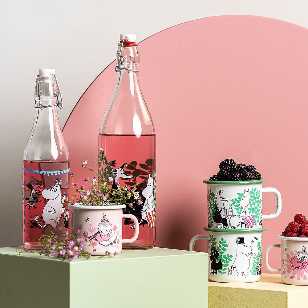 Muurla emalimukit ja lasipullot eri muumikuvituksilla, aseteltuna kukkien ja marjojen kanssa vaaleanvihreille alustoille