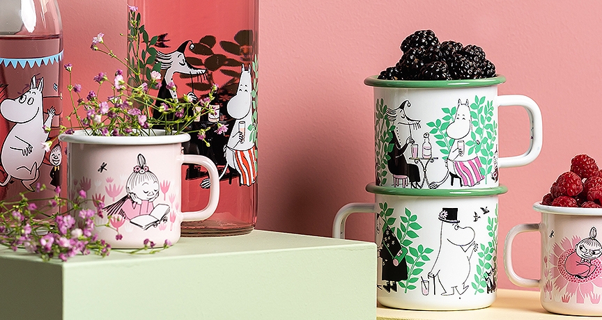 Muurla emalimukit ja lasipullot eri muumikuvituksilla, aseteltuna kukkien ja marjojen kanssa vaaleanvihreille alustoille