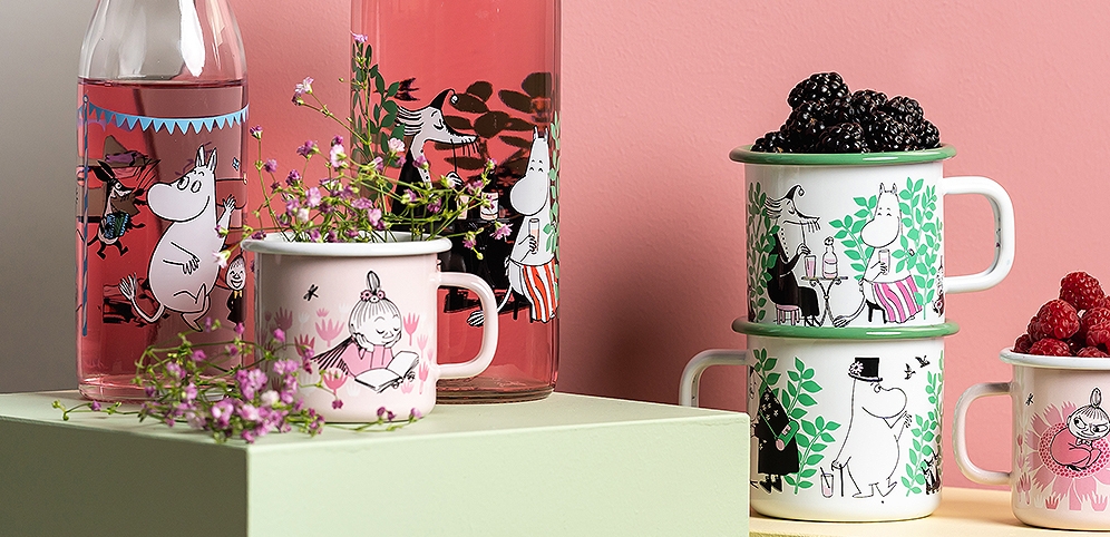 Muurla emalimukit ja lasipullot eri muumikuvituksilla, aseteltuna kukkien ja marjojen kanssa vaaleanvihreille alustoille