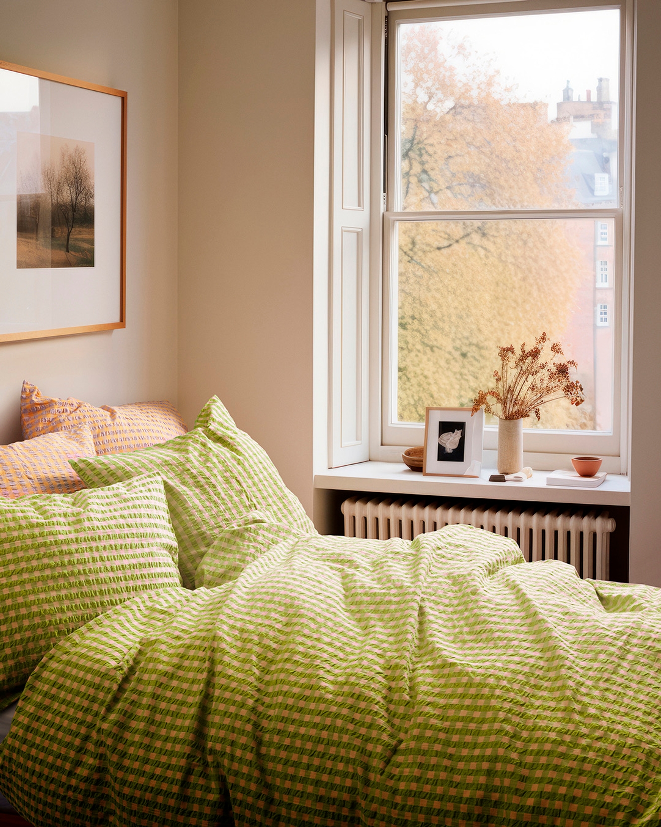A bed with green gingham bedding in a bright room with a window overlooking autumn trees and buildings.