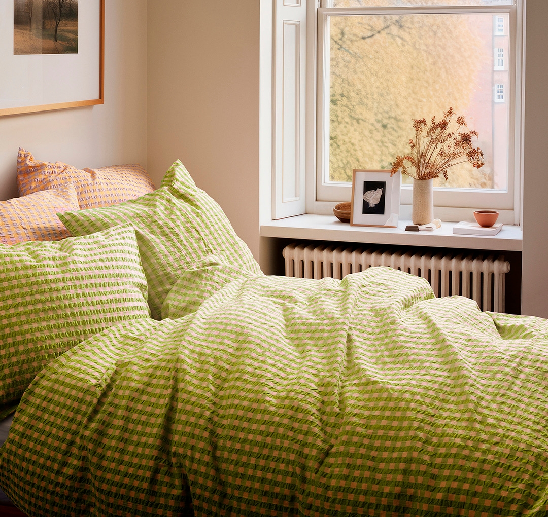 A bedroom with a bed made with green gingham bedding, a window overlooking autumn trees, and a framed picture on the wall.