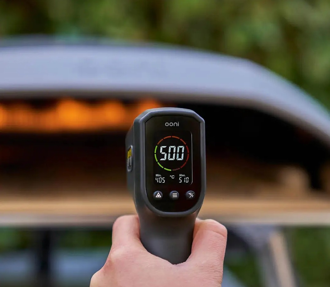 Hand som håller en Ooni IR-termometer som visar 500 °C framför en upphettad pizzaugn utomhus.