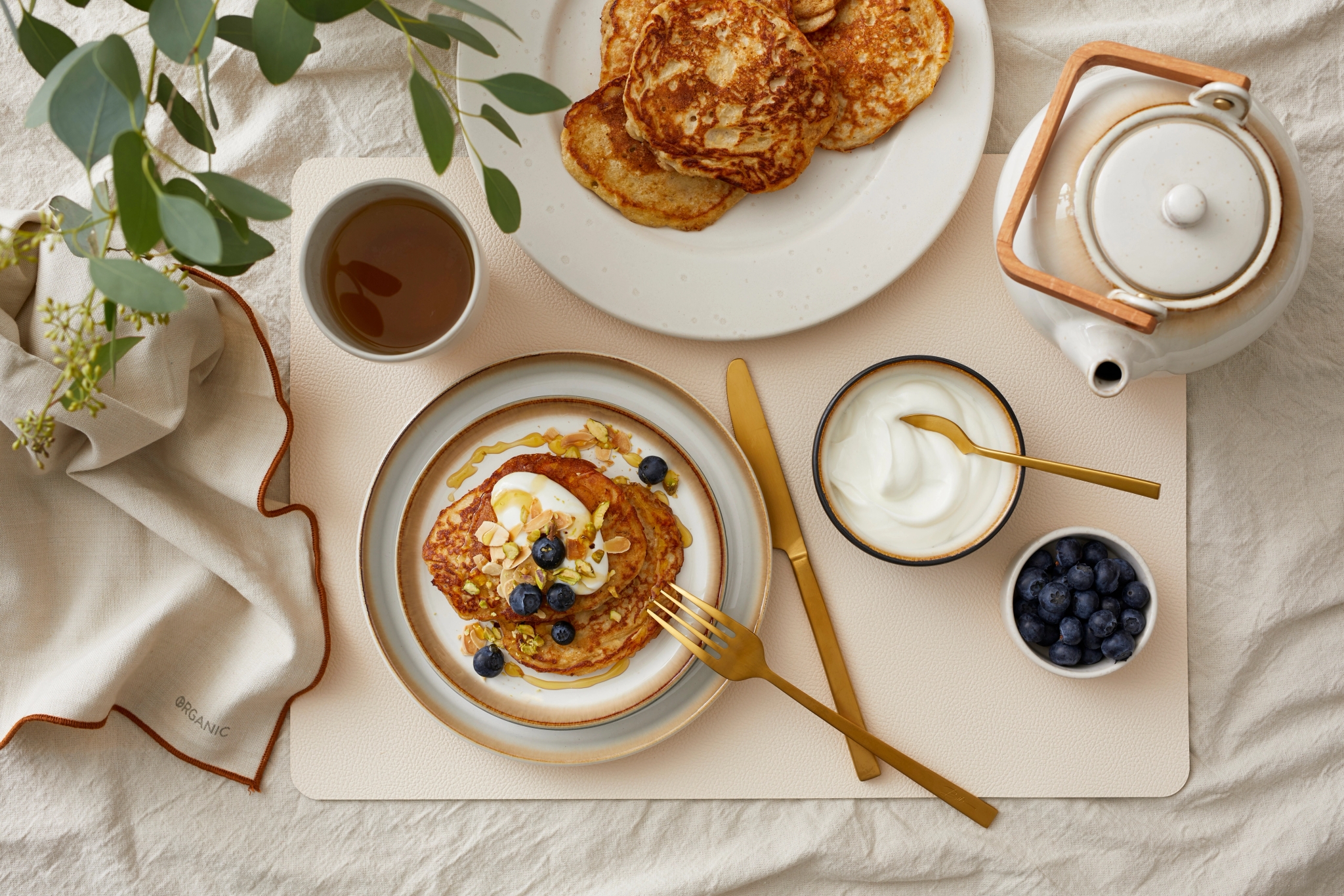 Frokostbord med pannekaker toppet med yoghurt, blåbær og nøtter, servert med te, bær og krem i lyse, nordiske omgivelser.