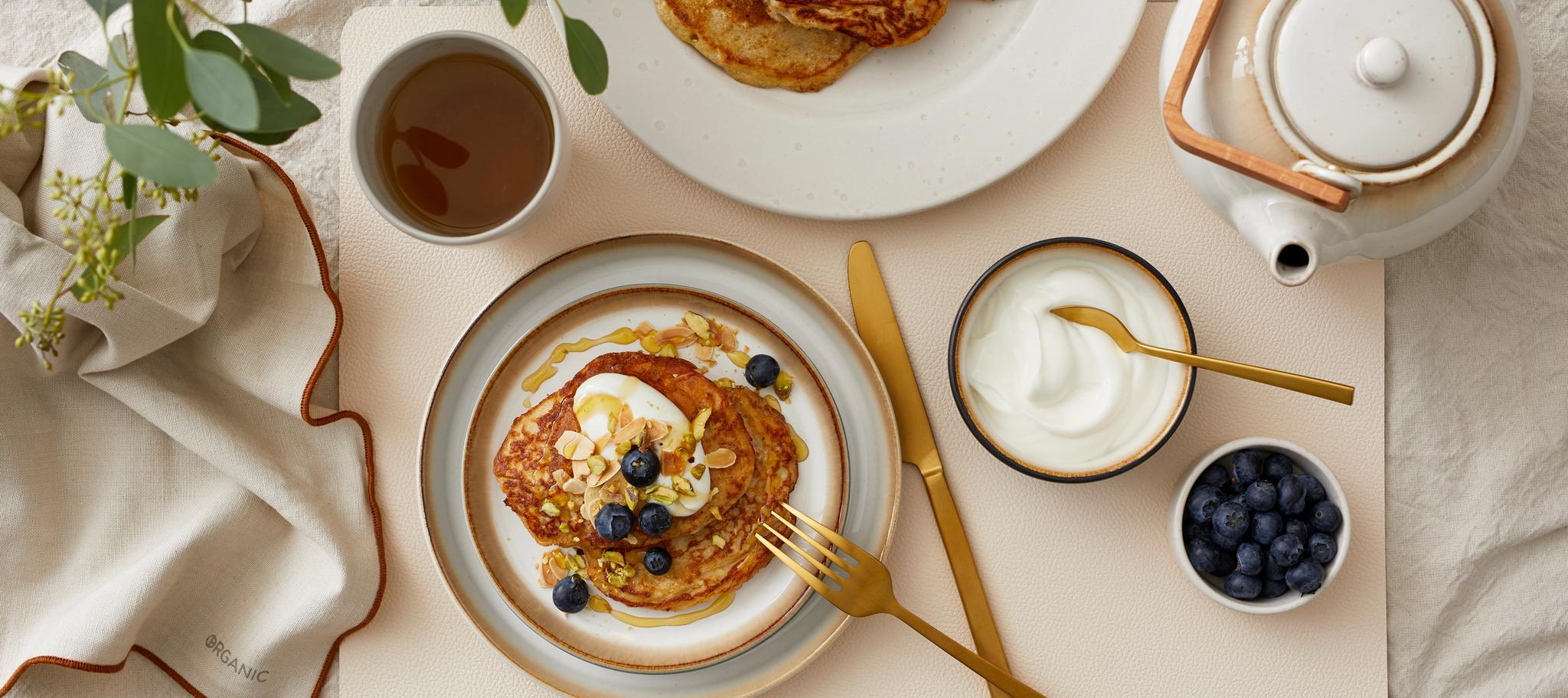 Lyxig frukost med pannkakor toppade med blåbär, yoghurt och nötter, serverade med te på ett dukat bord.