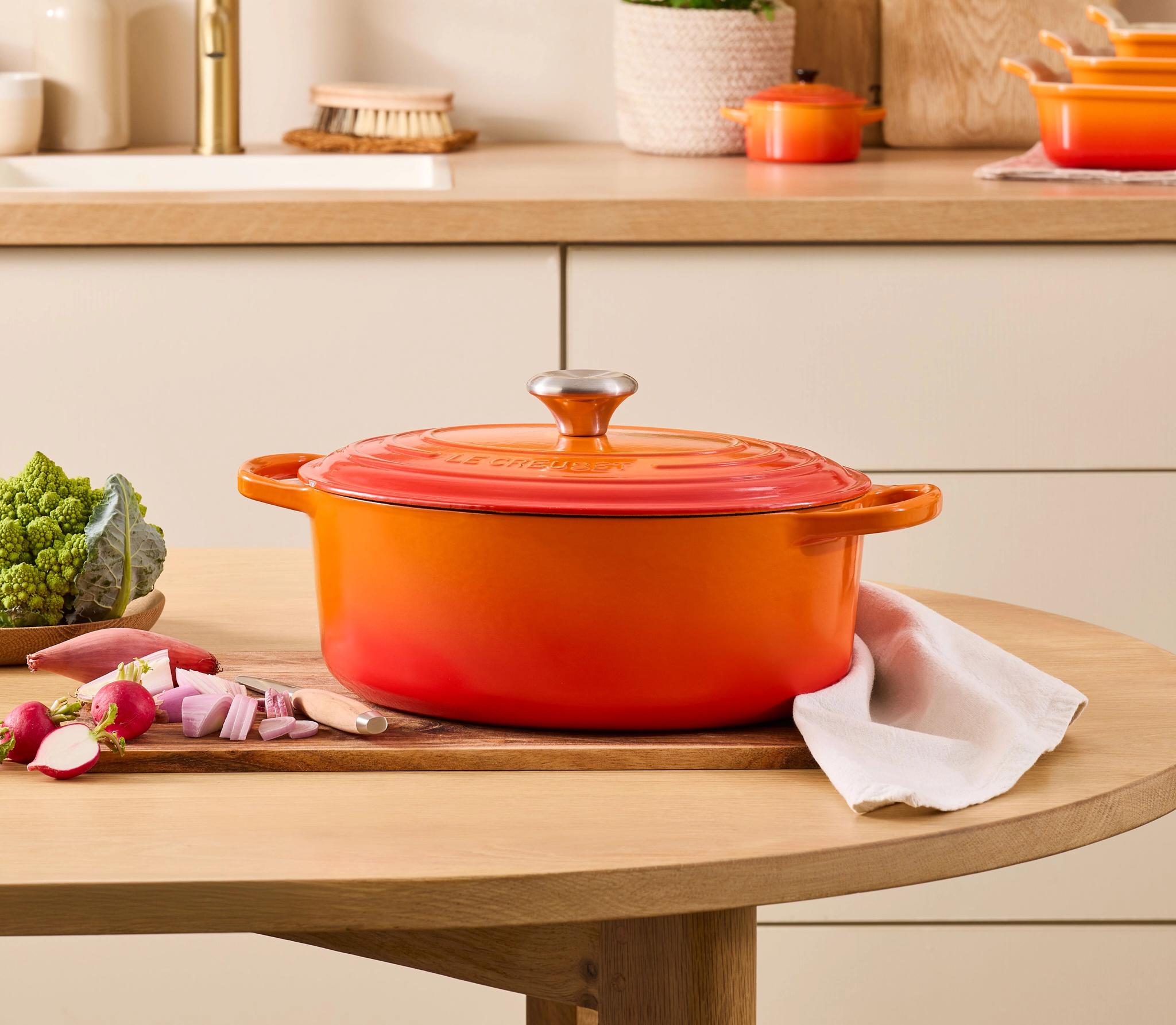 Oransje Le Creuset-gryte med lokk står på et kjøkkenbord med grønnsaker og skjærefjøl i et lyst, moderne kjøkken.