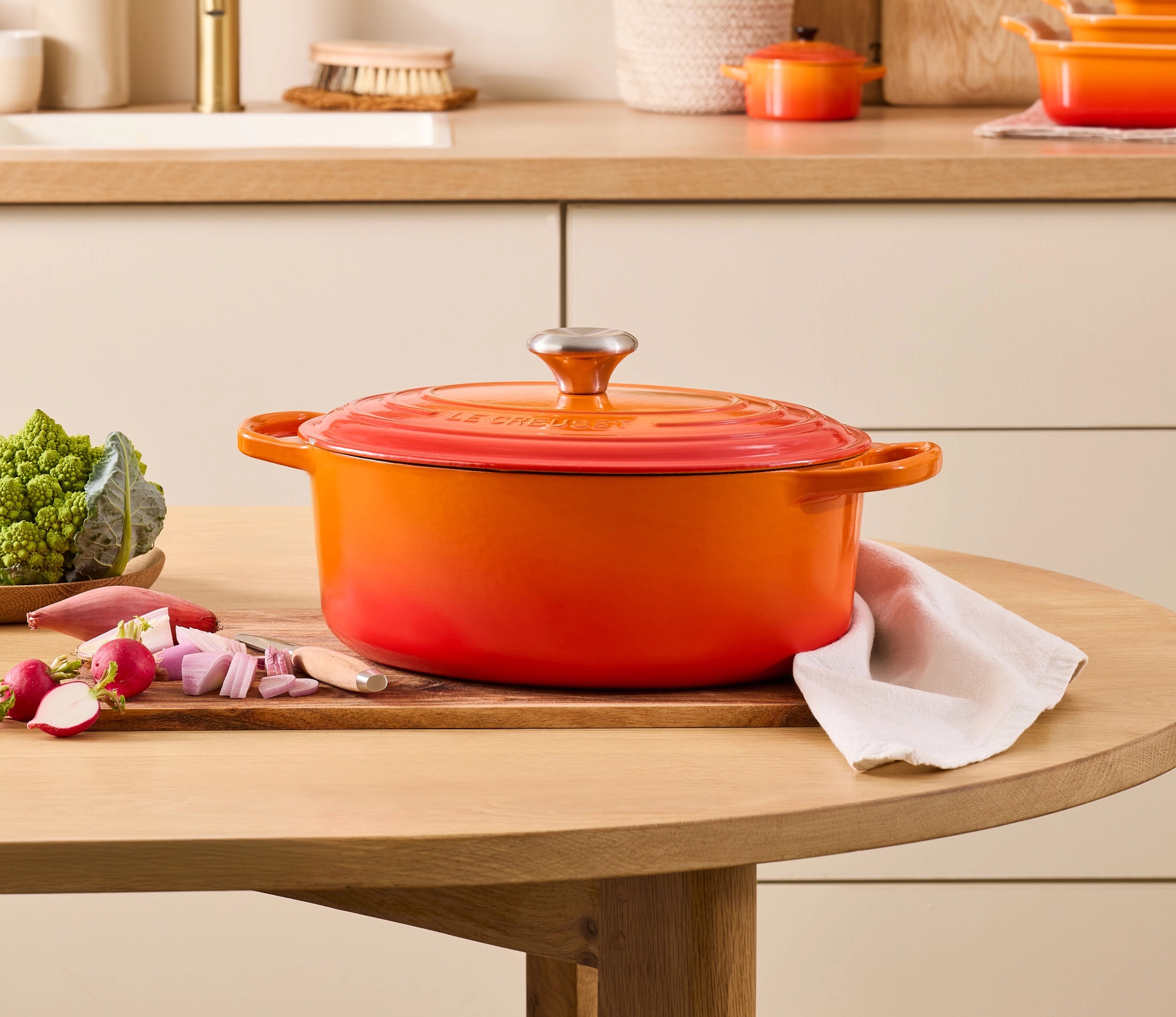 Oransje Le Creuset-gryte står på et kjøkkenbord med friske grønnsaker og lys, moderne kjøkkeninnredning i bakgrunnen.