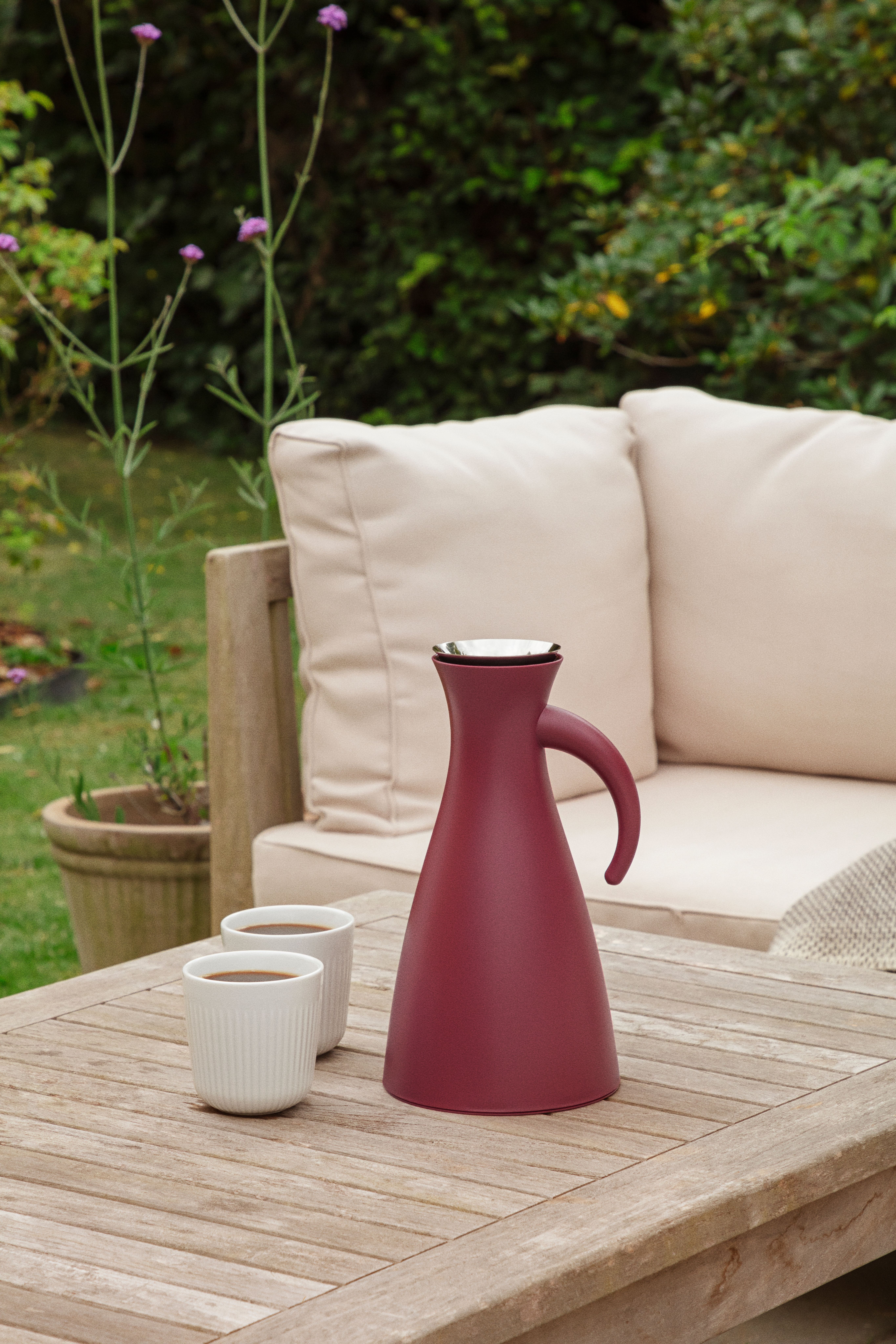 Burgunderrød termokanne og to kaffekopper står på et trebord på en uteplass med beige sofa og grønn hage i bakgrunnen.