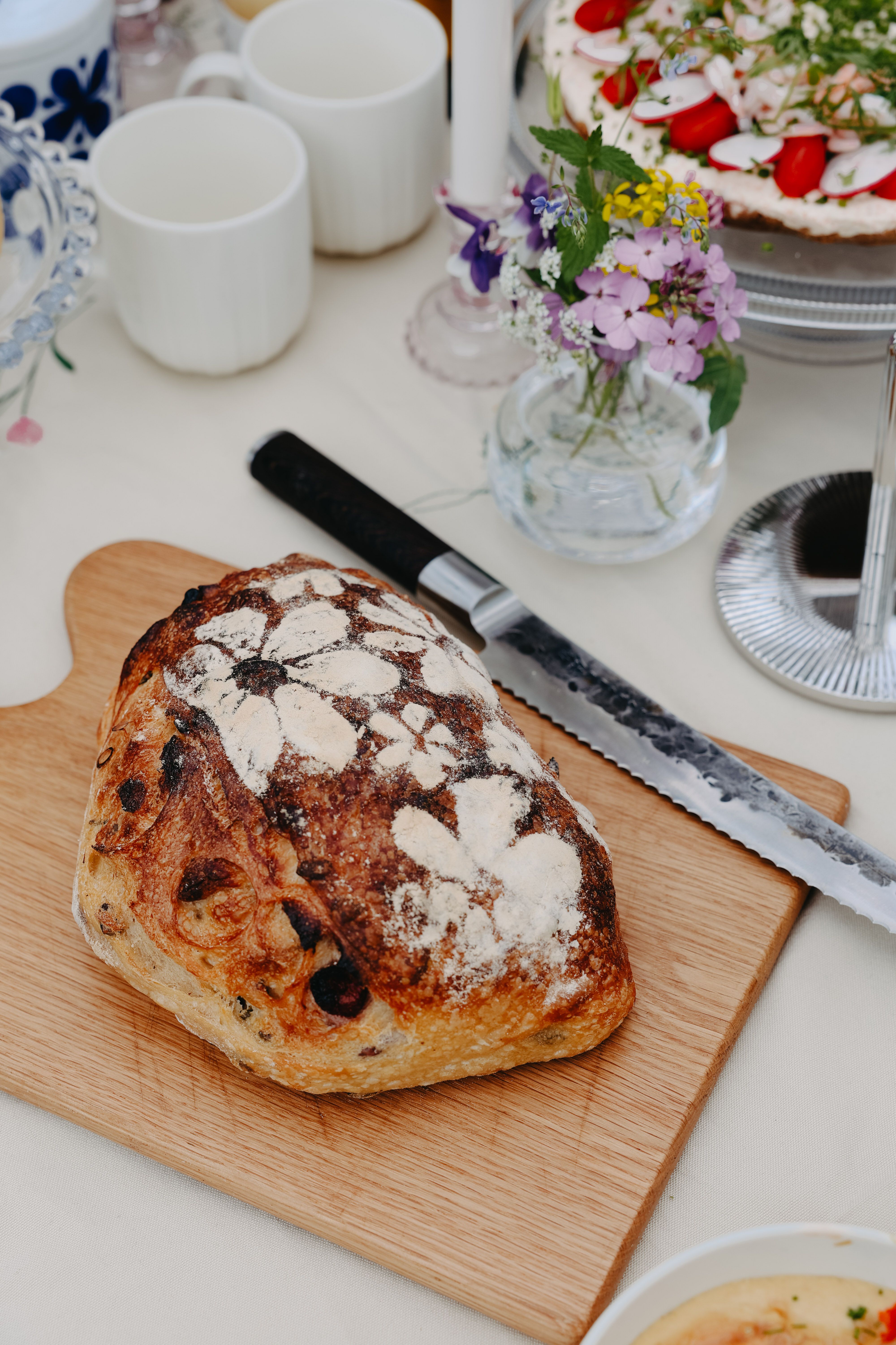 Nybakat surdegsbröd på skärbräda med brödkniv och somrig dukning i bakgrunden.