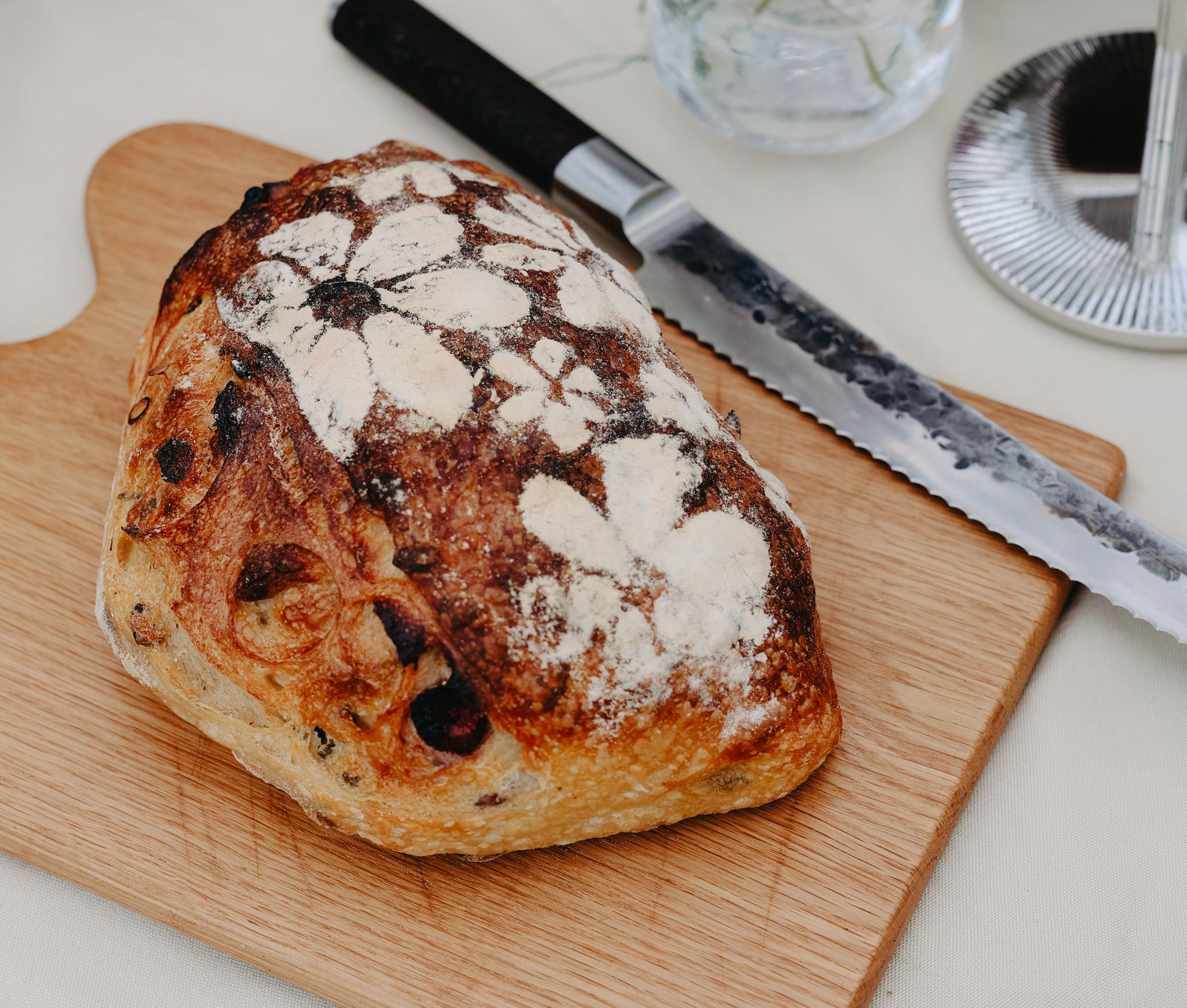 Nybakat surdegsbröd på skärbräda med brödkniv och somrig dukning i bakgrunden.