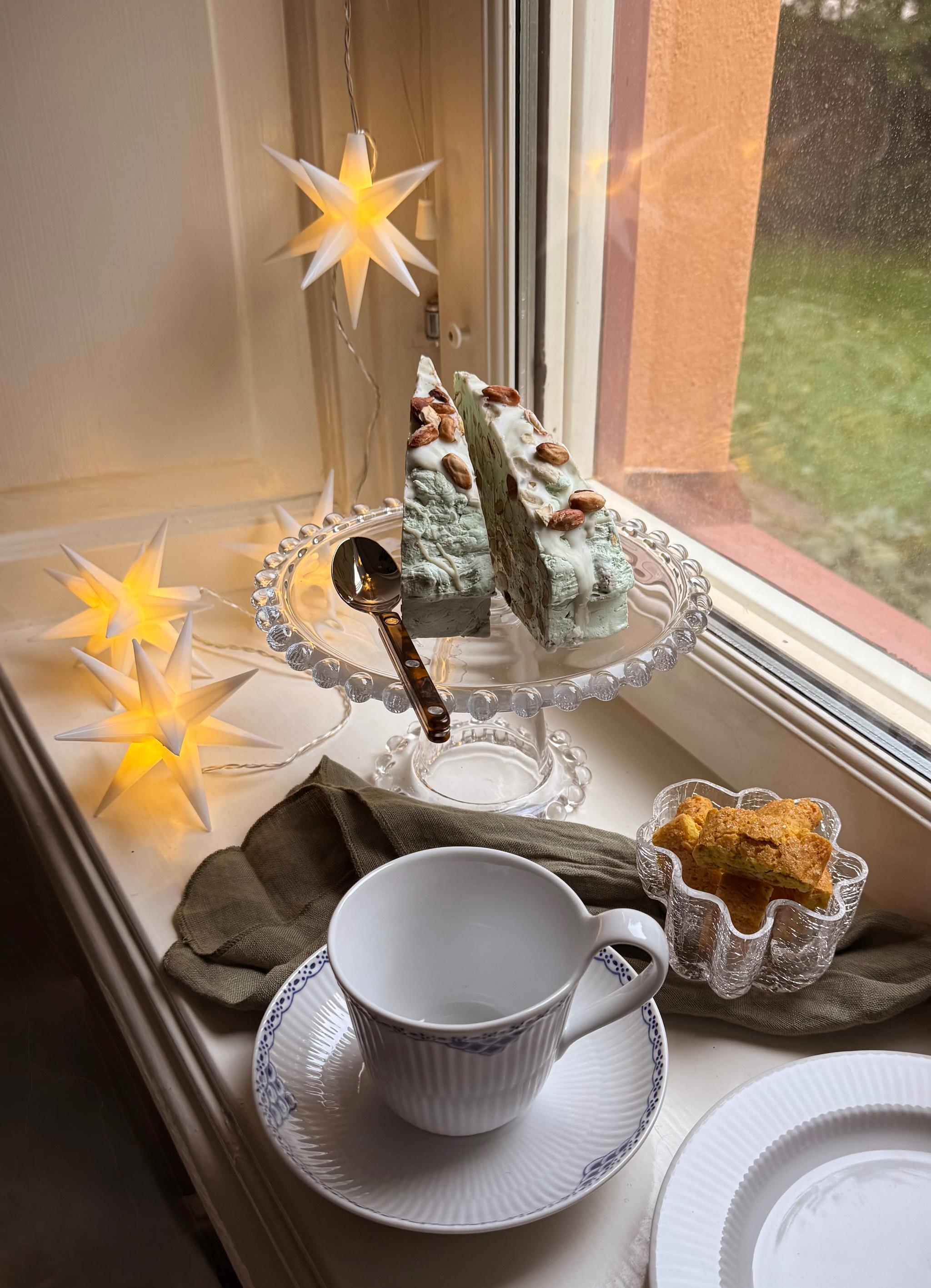 A festive window scene with glowing star lights, two slices of green almond-topped cake on a stand, a teacup, and cookies.