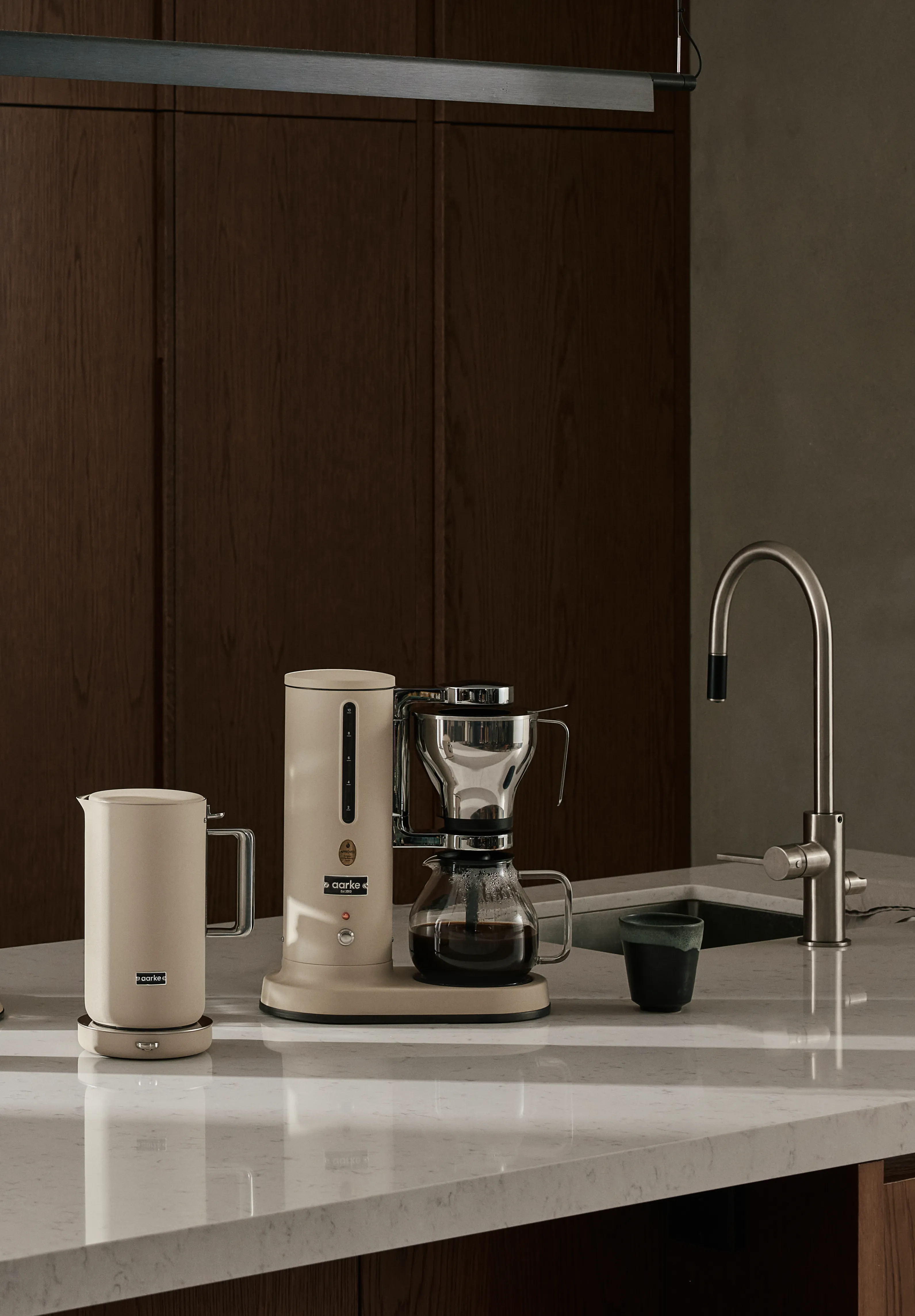 En beige kaffebryggare och matchande vattenkokare på en vit marmorbänkskiva med ett handfat, en kran och en mörk mugg, mot mörka träskåp.