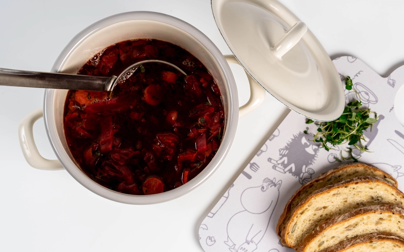 Punainen keitto kattilassa kauhan kanssa, vieressä siivutettua leipää ja yrttejä kuviollisella valkoisella leikkuulaudalla.