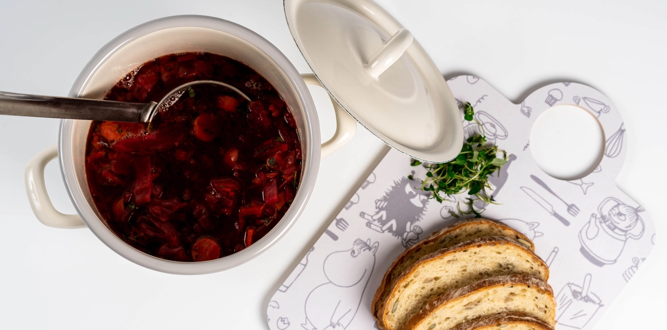 Punainen keitto kattilassa kauhan kanssa, vieressä siivutettua leipää ja yrttejä kuviollisella valkoisella leikkuulaudalla.