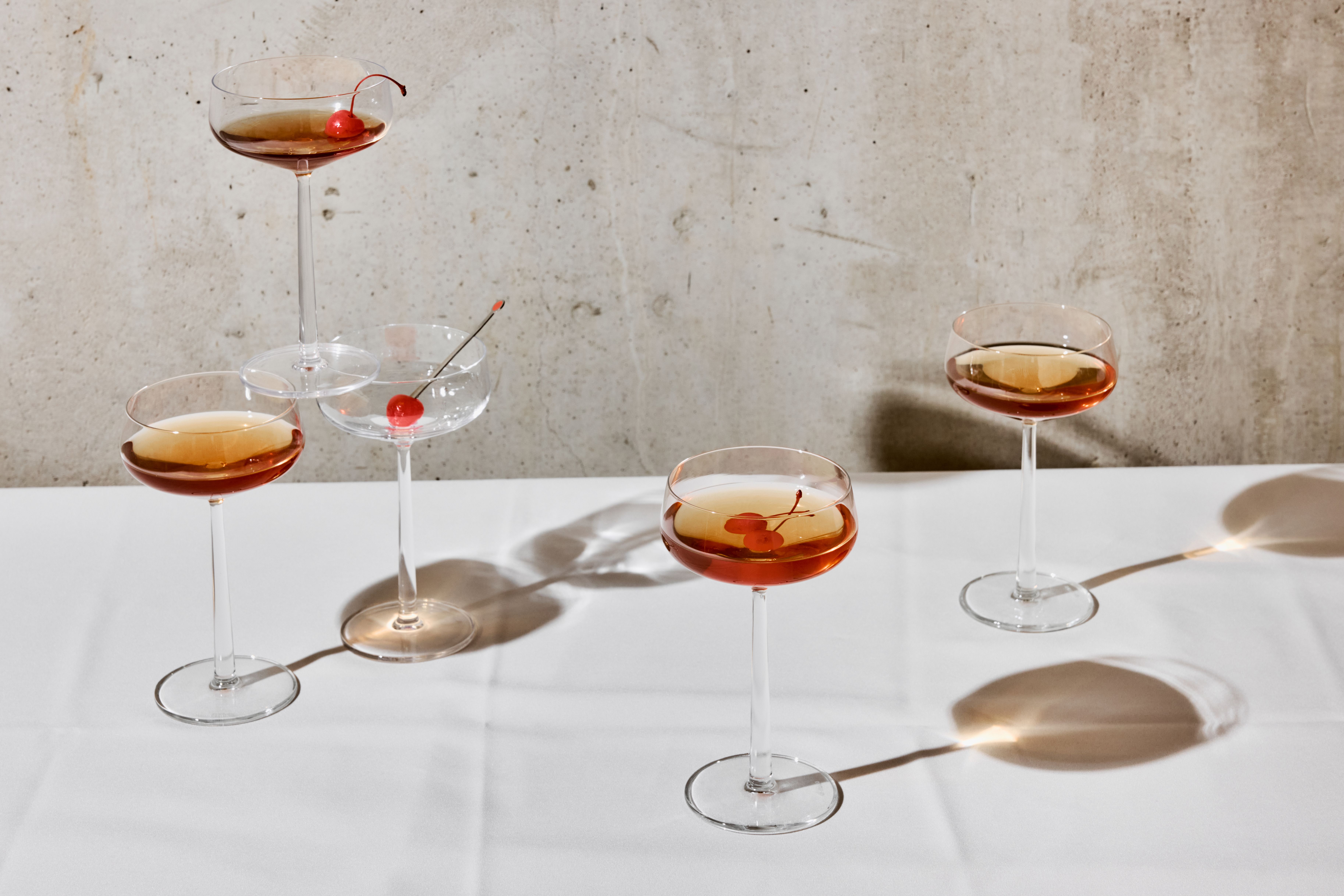 Viisi kuppilasia, joissa on meripihkanvärisiä cocktaileja ja kirsikoita, heittävät pitkiä varjoja valkoiselle pöydälle betoniseinää vasten.