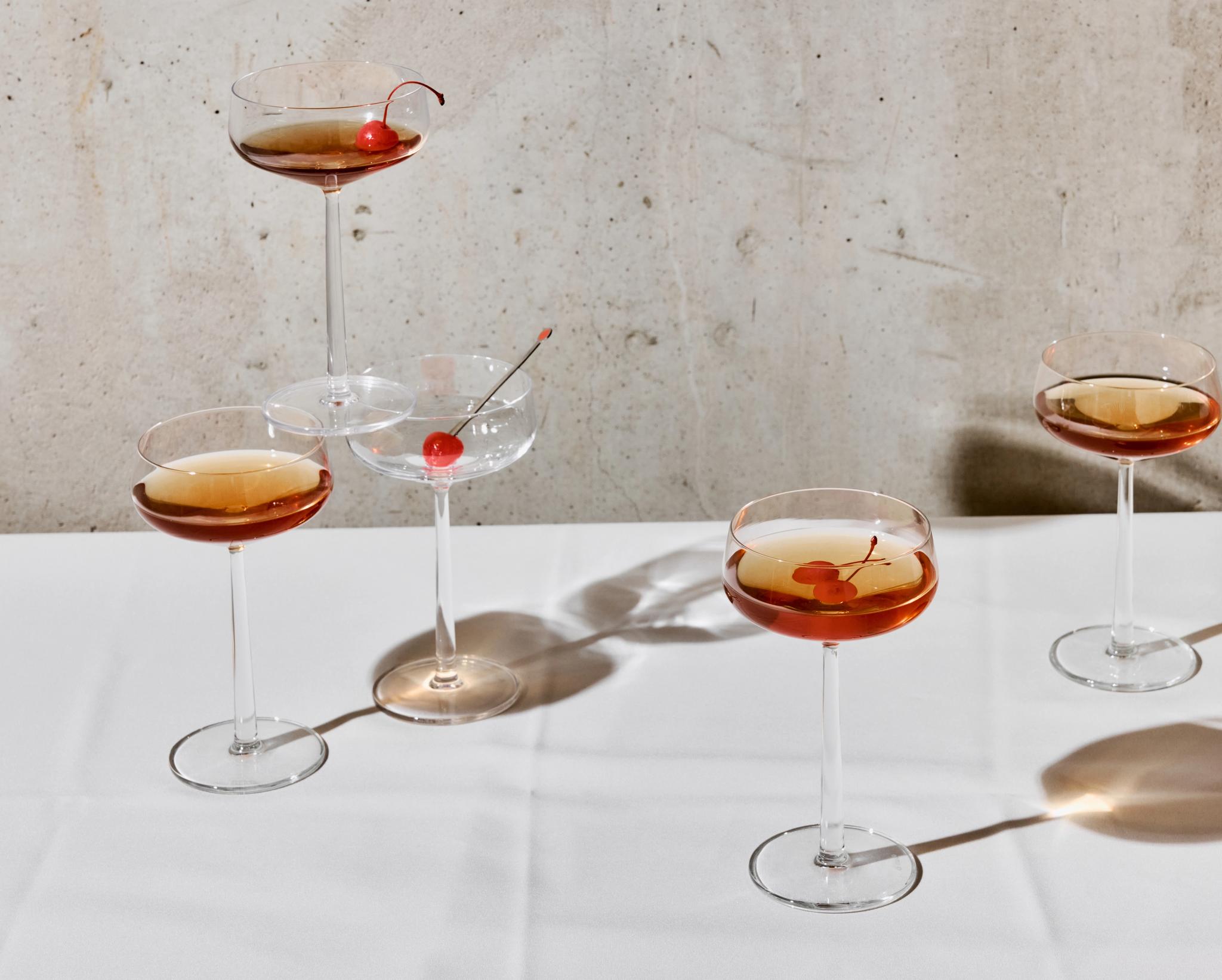 Useita tyylikkäitä cocktail-laseja, joissa on ruskeasävyisiä juomia ja kirsikoita, valkoisella pöytäliinalla betoniseinän edessä.