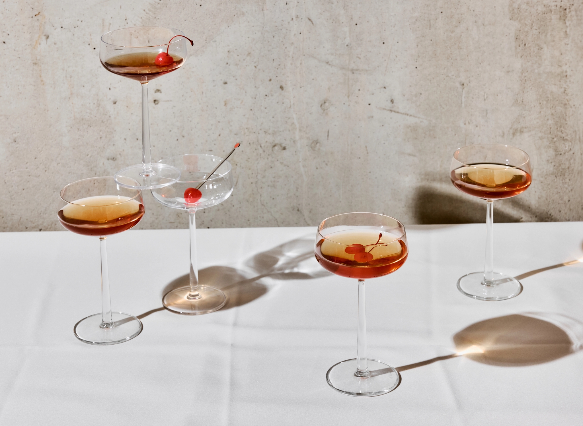 Tyylikkäät cocktail-lasit, joissa on ruskeasävyisiä juomia ja kirsikoita, seisovat valkoisella pöytäliinalla betoniseinän edessä.