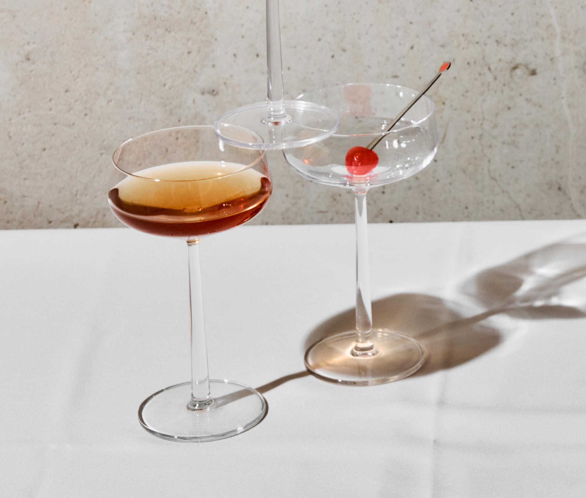 Viisi kuppilasia, joissa on meripihkanvärisiä cocktaileja ja kirsikoita, heittävät pitkiä varjoja valkoiselle pöydälle betoniseinää vasten.